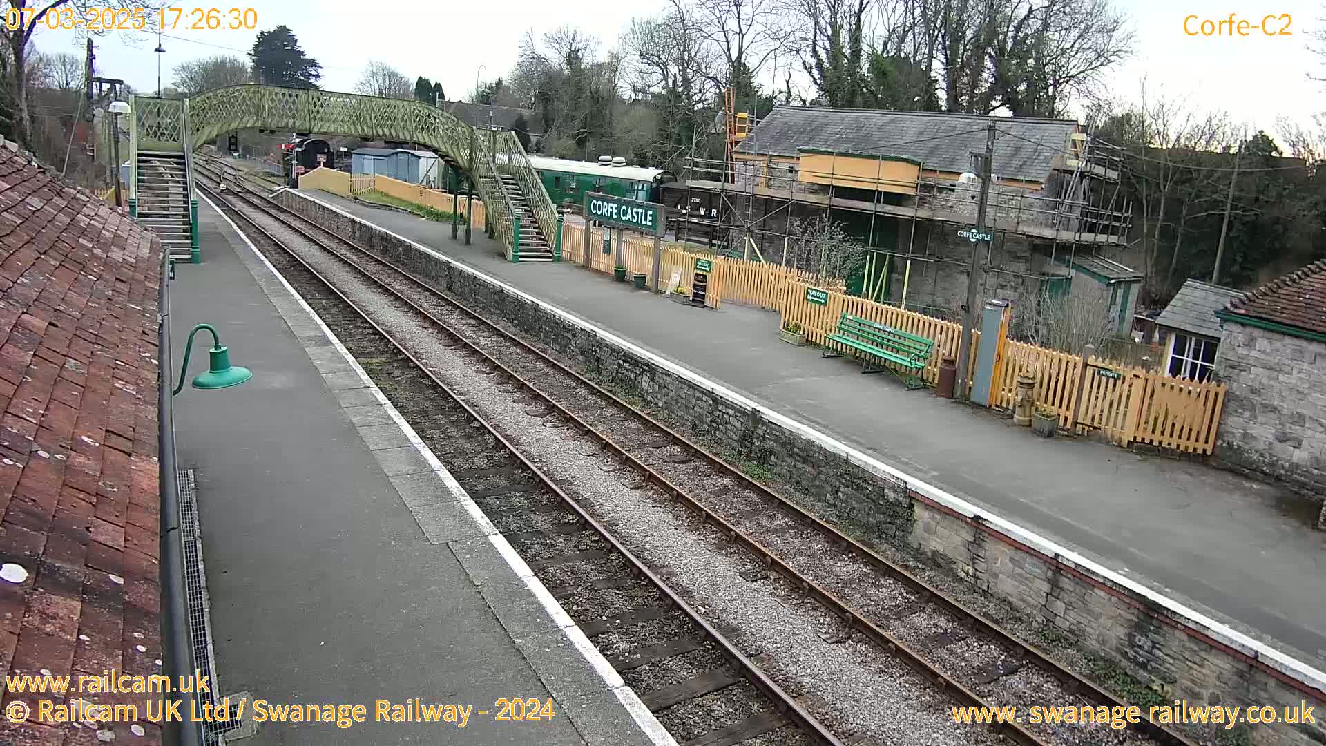 A train station with a railway track, a pedestrian bridge, and several buildings under an overcast sky.