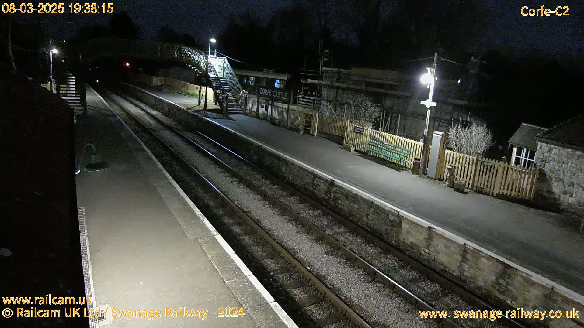 A nighttime view of a train station platform with two train tracks, a pedestrian bridge, and some buildings under dark skies.