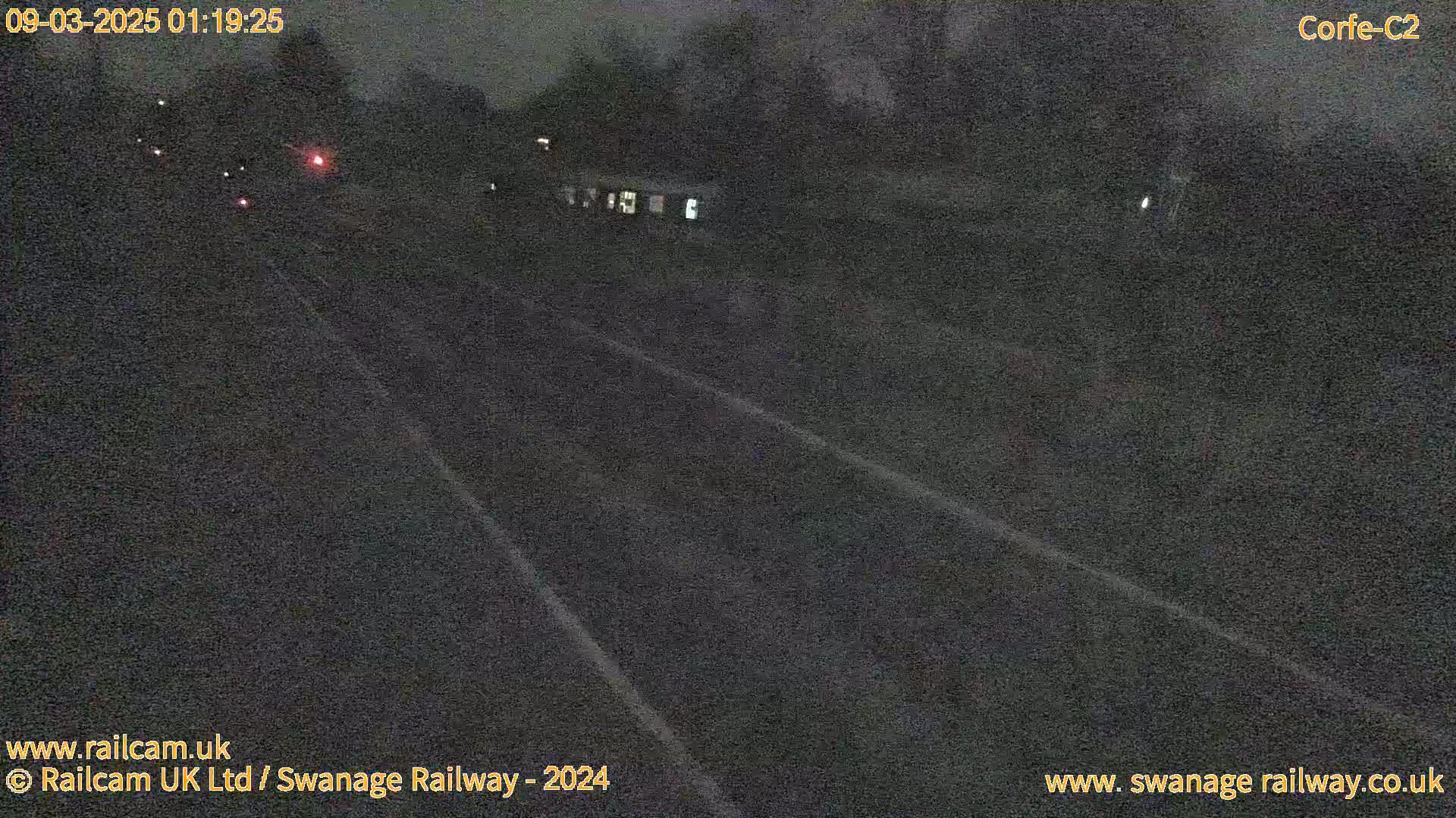 A dark nighttime view of a road or railway track with a train partially visible in the distance under a cloudy sky.