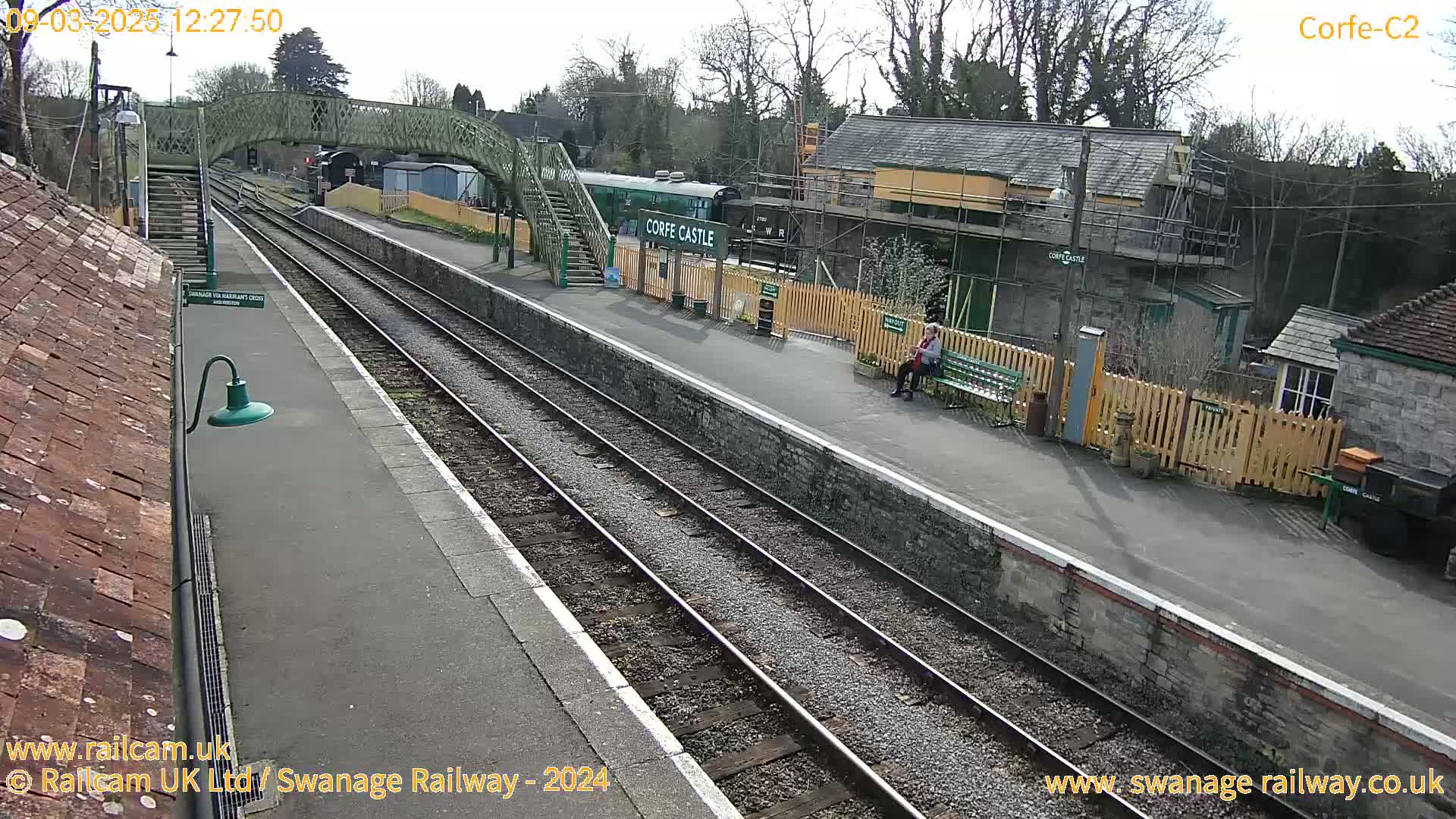 A train station platform with train tracks, a railway car, a pedestrian bridge, and a person sitting on a bench on a sunny day.