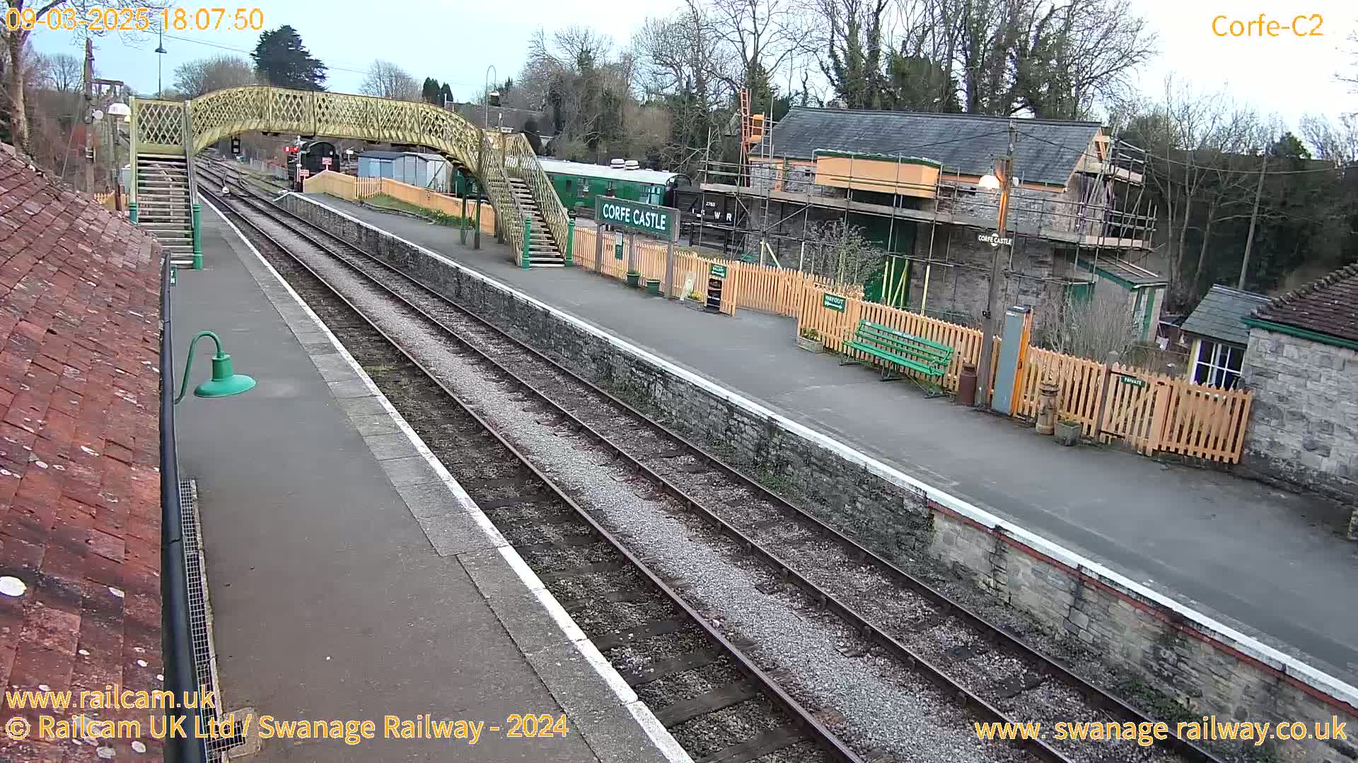 A train station with a wooden footbridge, railway tracks, and a building under construction is visible on an overcast day.