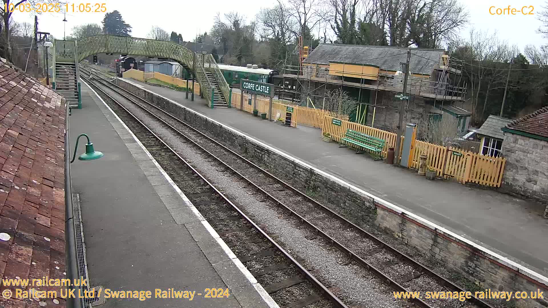 A railway station platform with train tracks, a green train, and buildings under a partly cloudy sky.