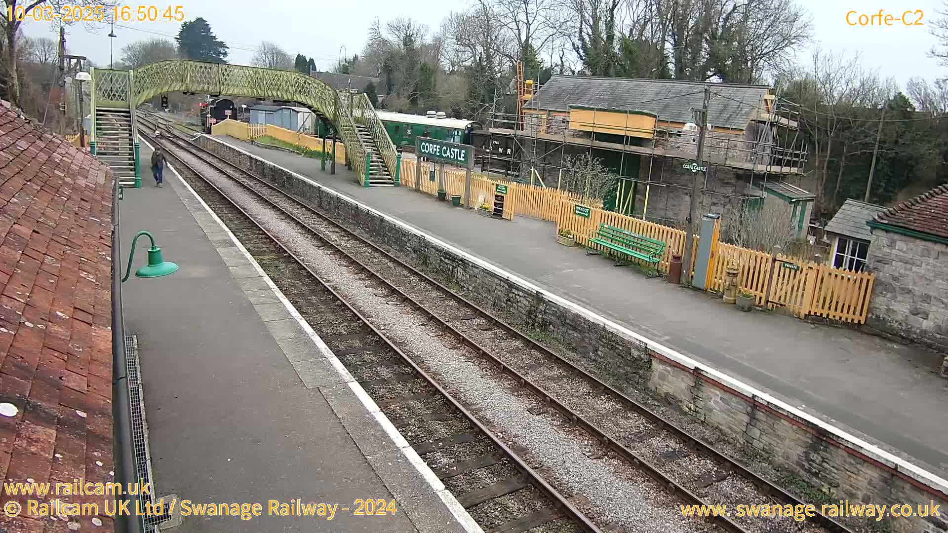 A person walks on a train platform next to railway tracks and buildings under a partly cloudy sky.
