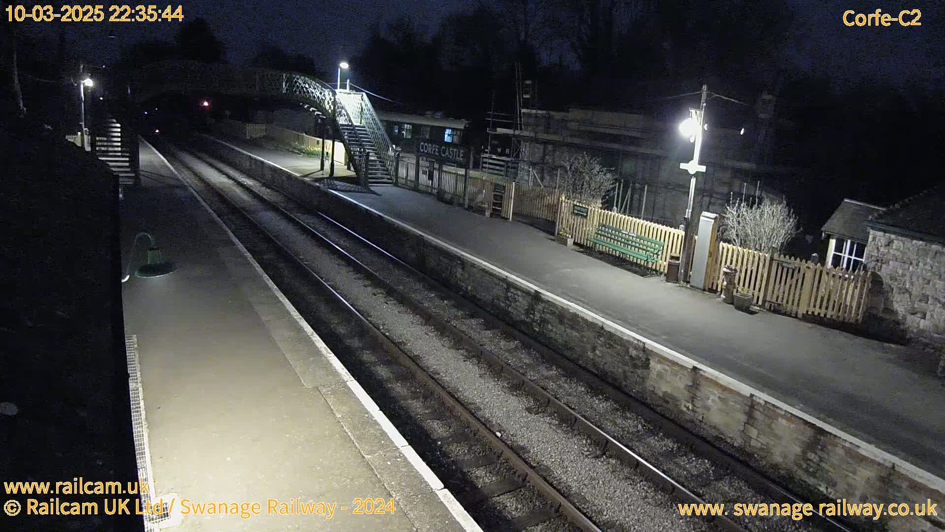 A nighttime view of a deserted train station platform with two sets of train tracks, a pedestrian bridge, and a few lights illuminating the scene.