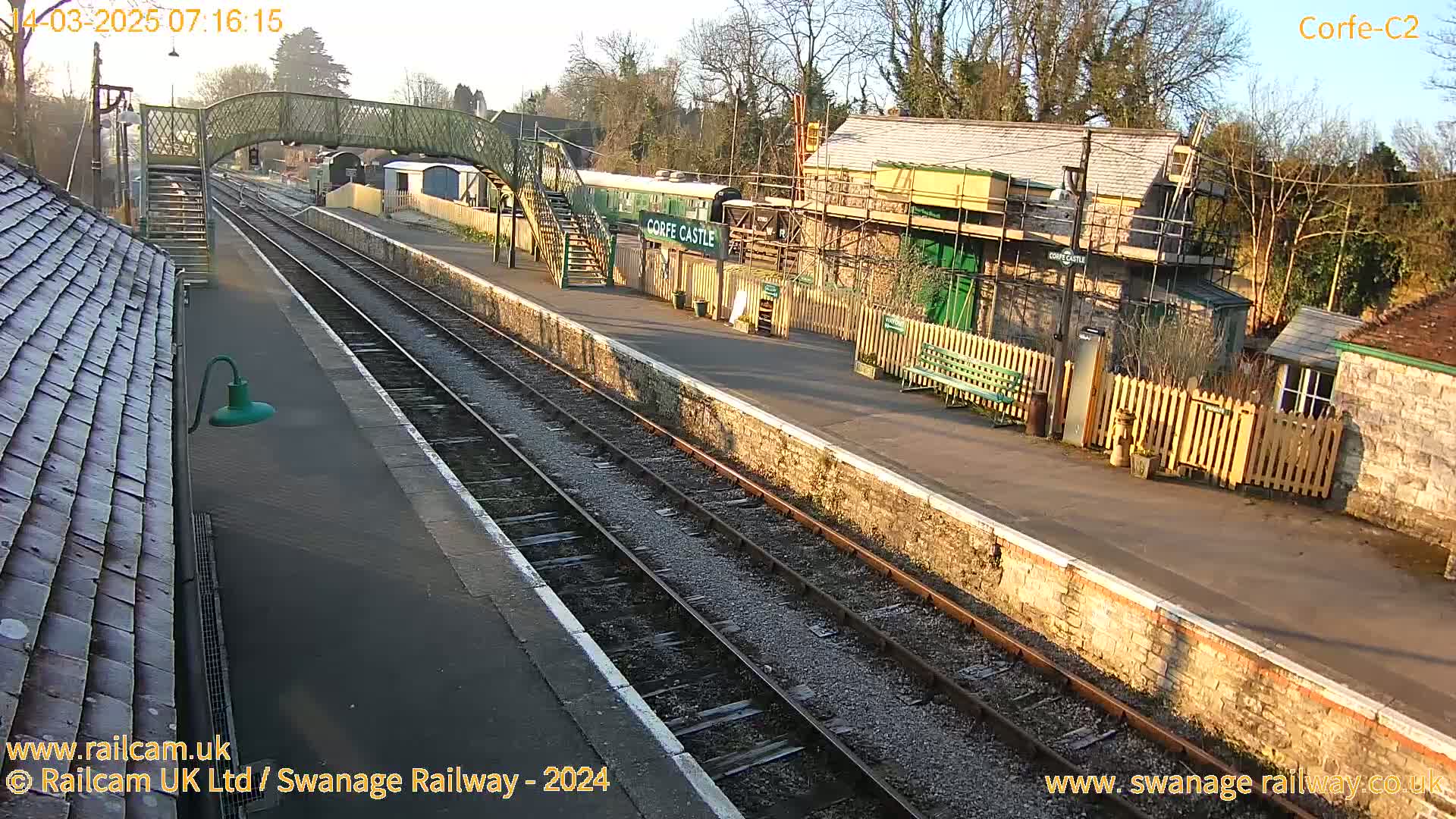 A sunny morning view of a train station with multiple tracks, a pedestrian bridge, and buildings undergoing construction or renovation.