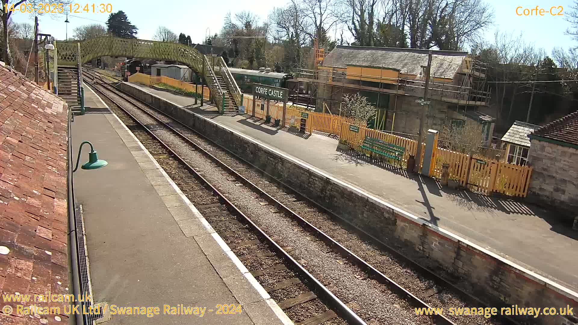 A sunny day at a train station with a railway track, a platform, and buildings under construction, including a train stopped at the platform.