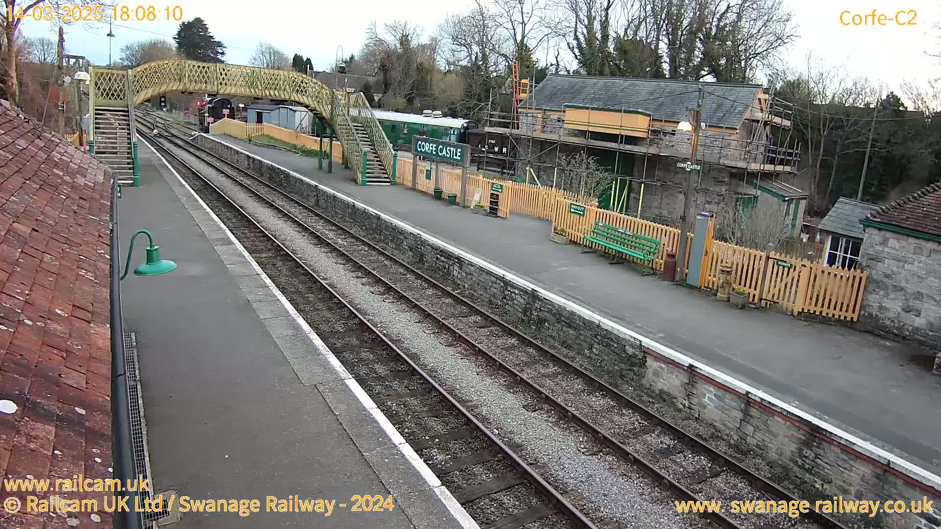 A train station with two railway tracks, a wooden footbridge, and a building under construction is seen on an overcast day.