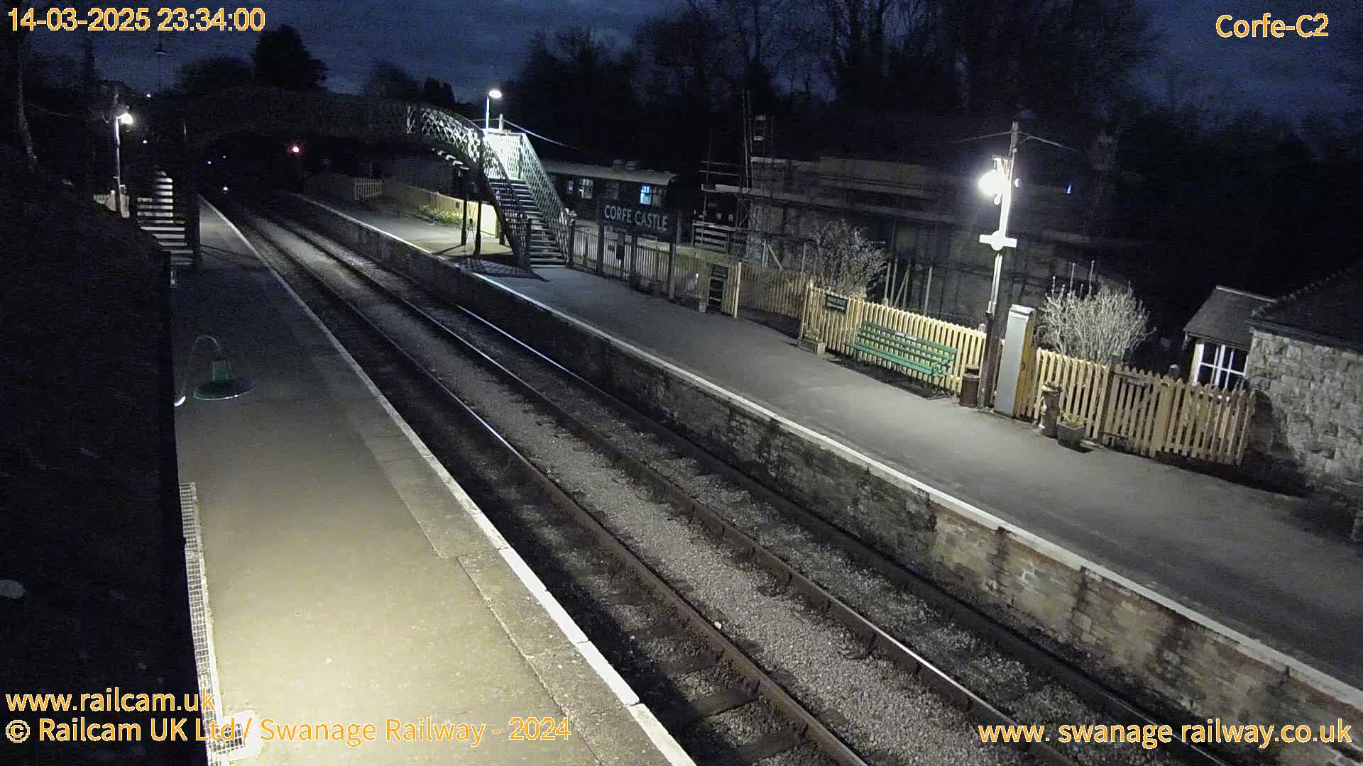 An empty train platform at night is illuminated by artificial lights, with train tracks extending into the darkness.
