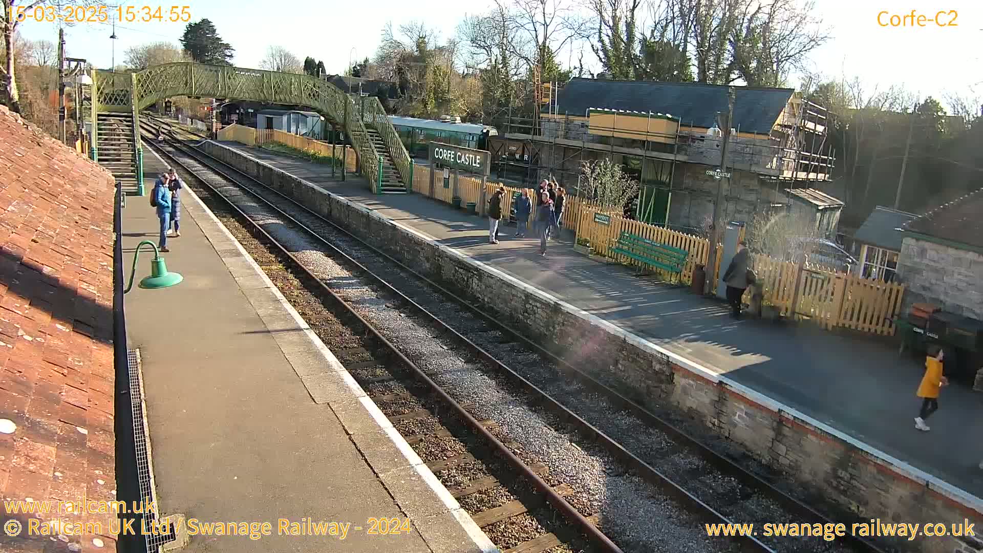 A sunny day at a train station shows several people gathered on the platform near a railway car and building under construction.