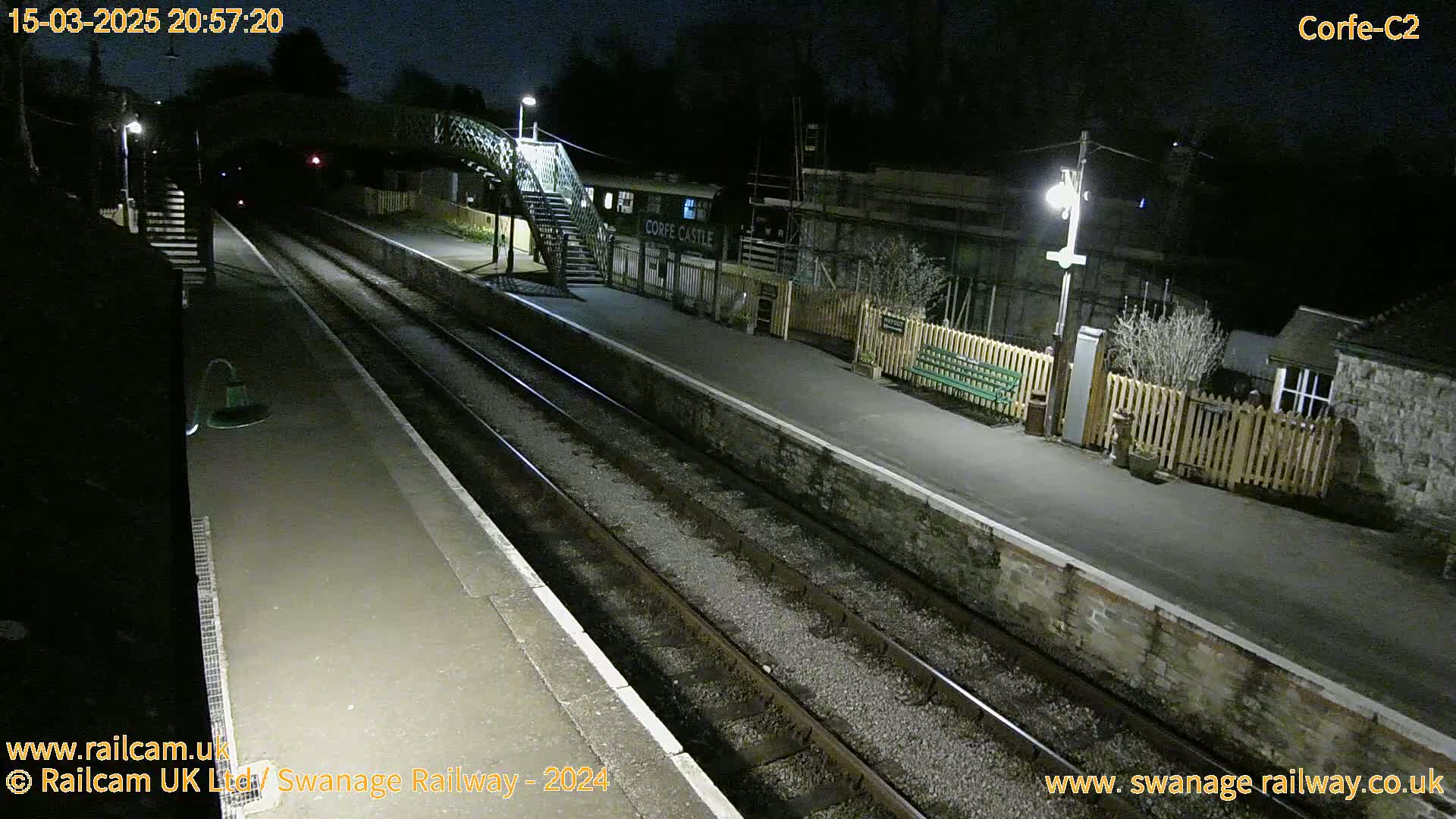 A nighttime view of a train station platform with two sets of train tracks, a pedestrian bridge, and a building.