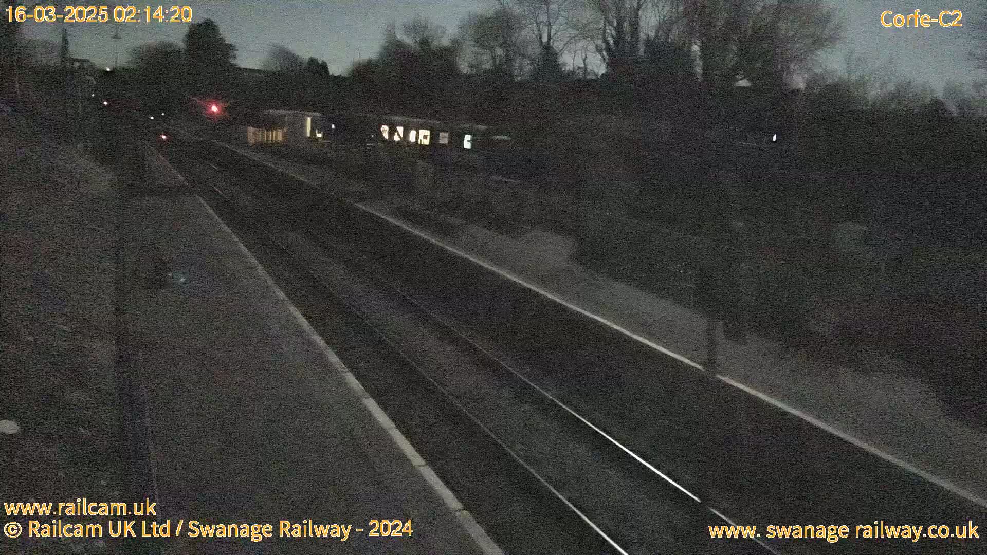 A nighttime view of a train track and platform, under dark, possibly overcast skies.