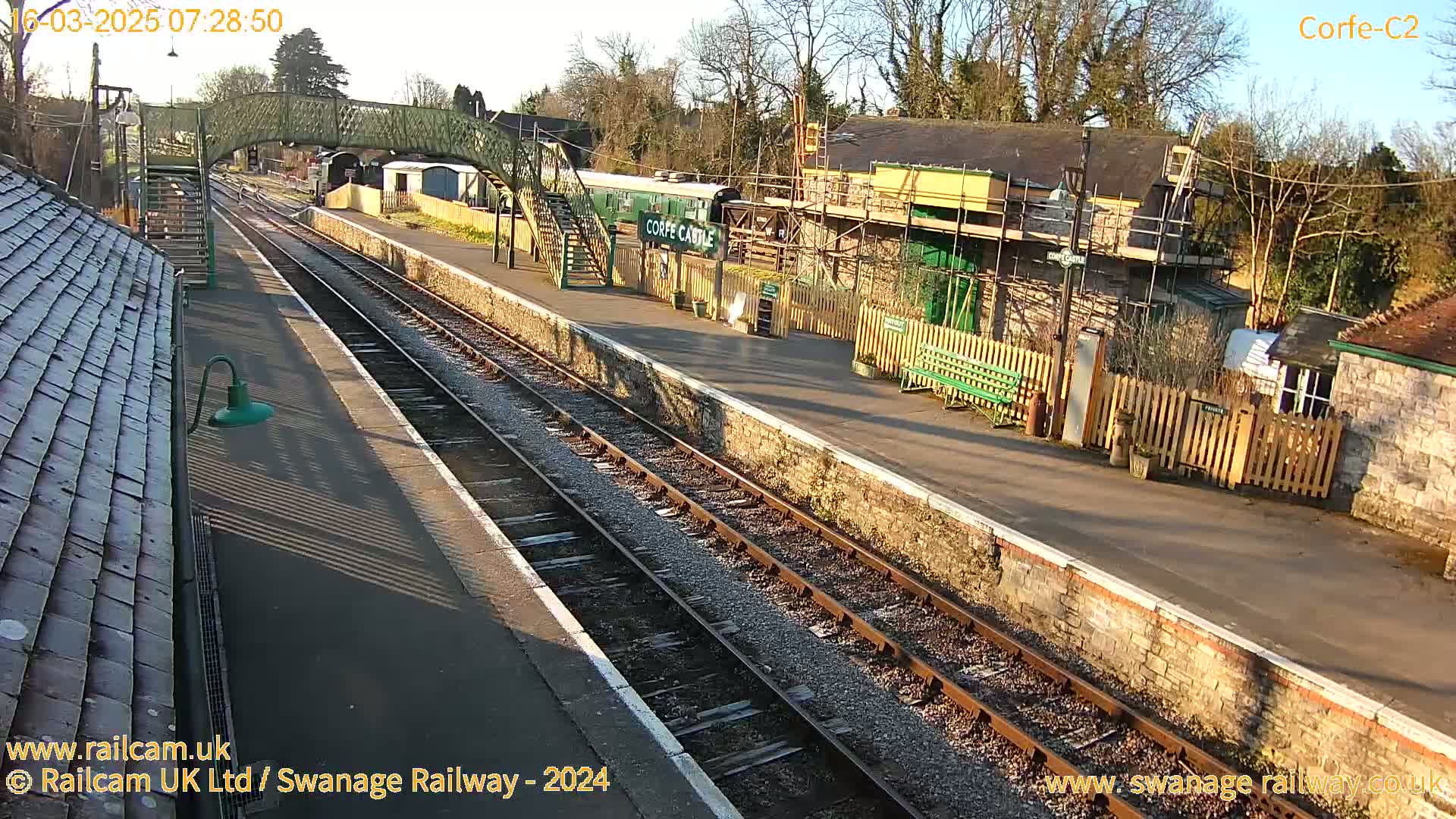 A sunny view of a train station with two parallel tracks, a railway carriage, a footbridge, and some buildings undergoing renovations.