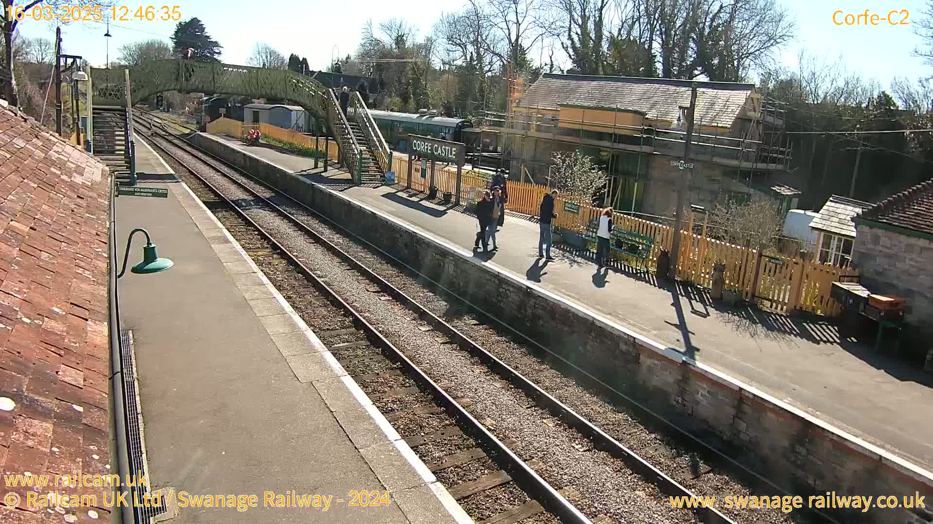 A sunny day at a train station shows a train stopped at the platform with several people standing nearby, alongside a footbridge and various buildings.