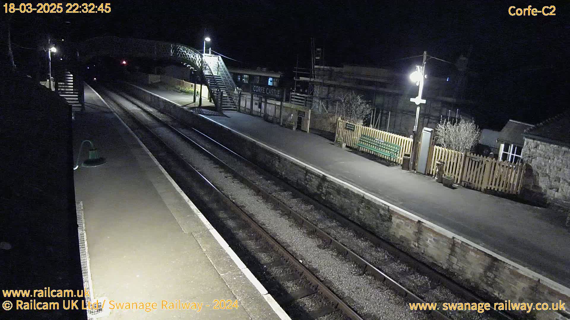 A nighttime view of a deserted train station platform with two sets of train tracks, a footbridge, and a building.