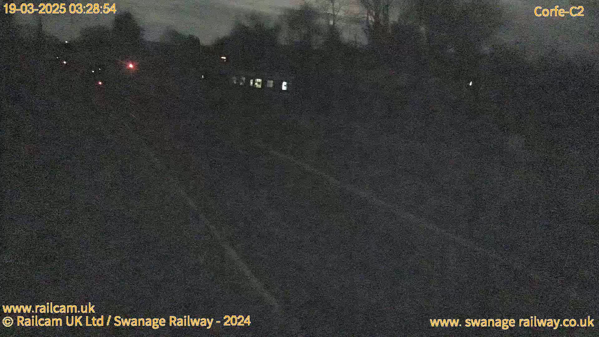 A dark, nighttime view of a road or railway track with sparse, distant lights and trees.
