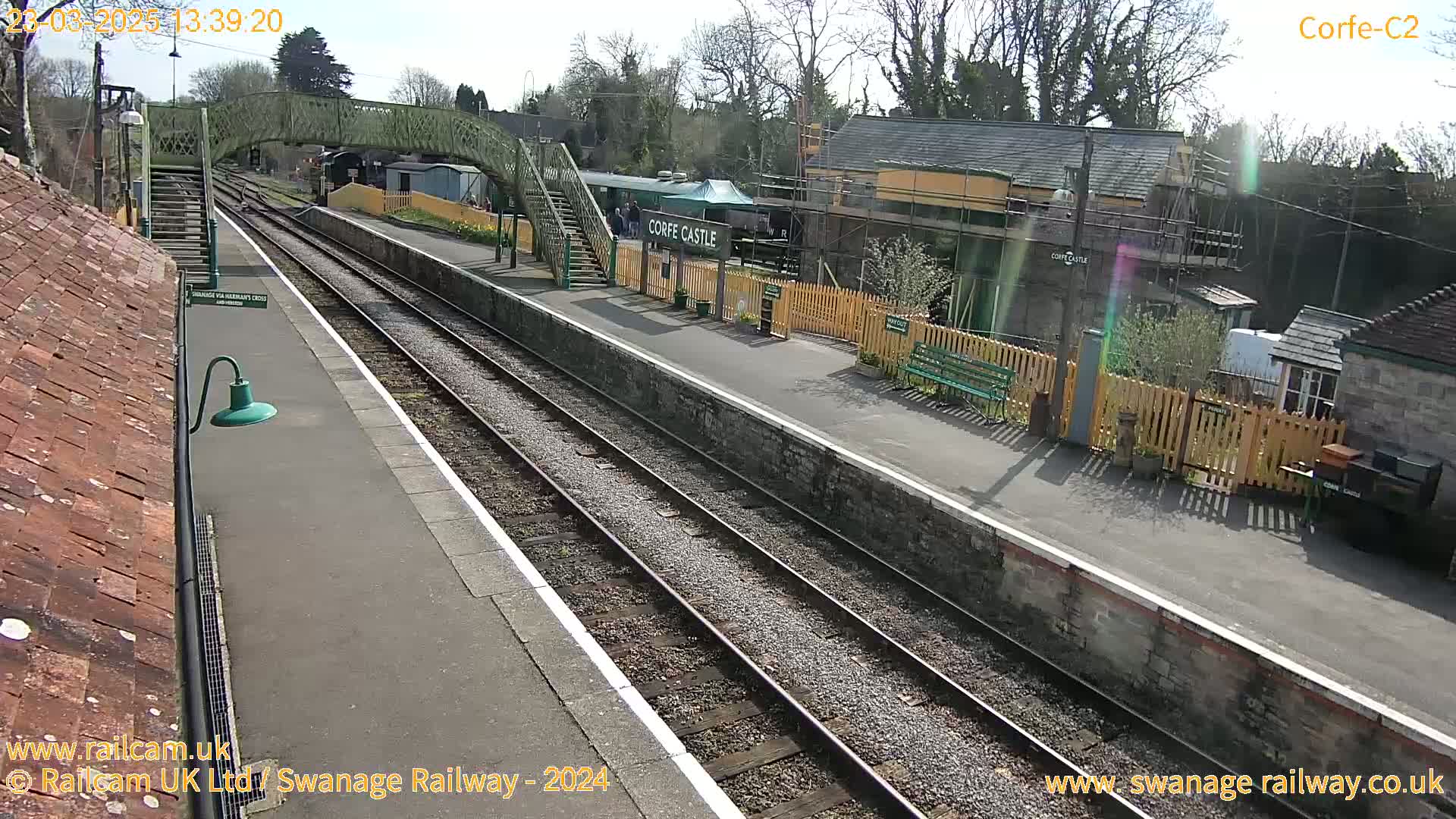 A sunny view of a train station platform with two train tracks, a footbridge, and several buildings, some under construction.
