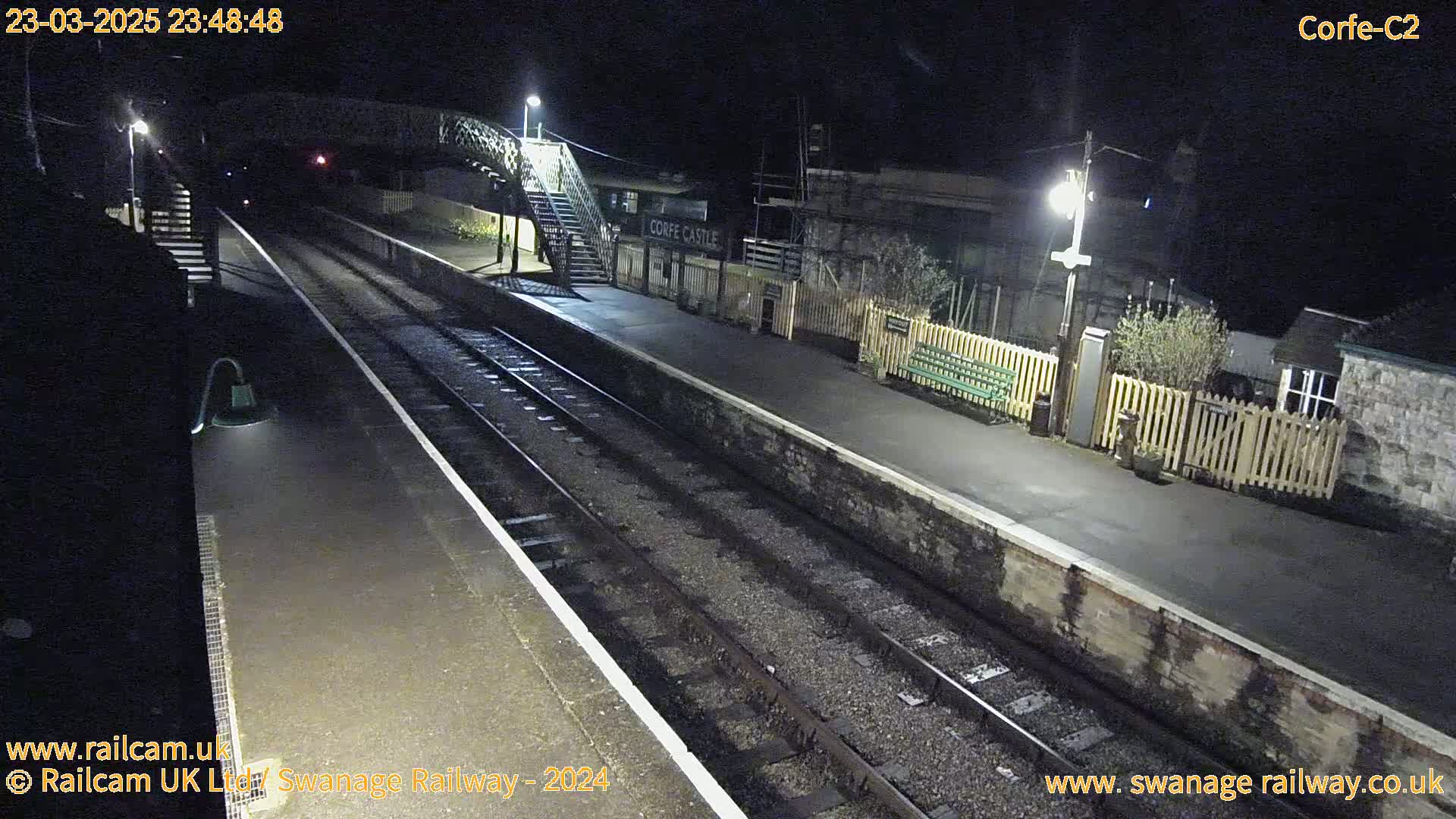 A nighttime view of a deserted train station platform with two sets of train tracks, a footbridge, and a low wooden fence.