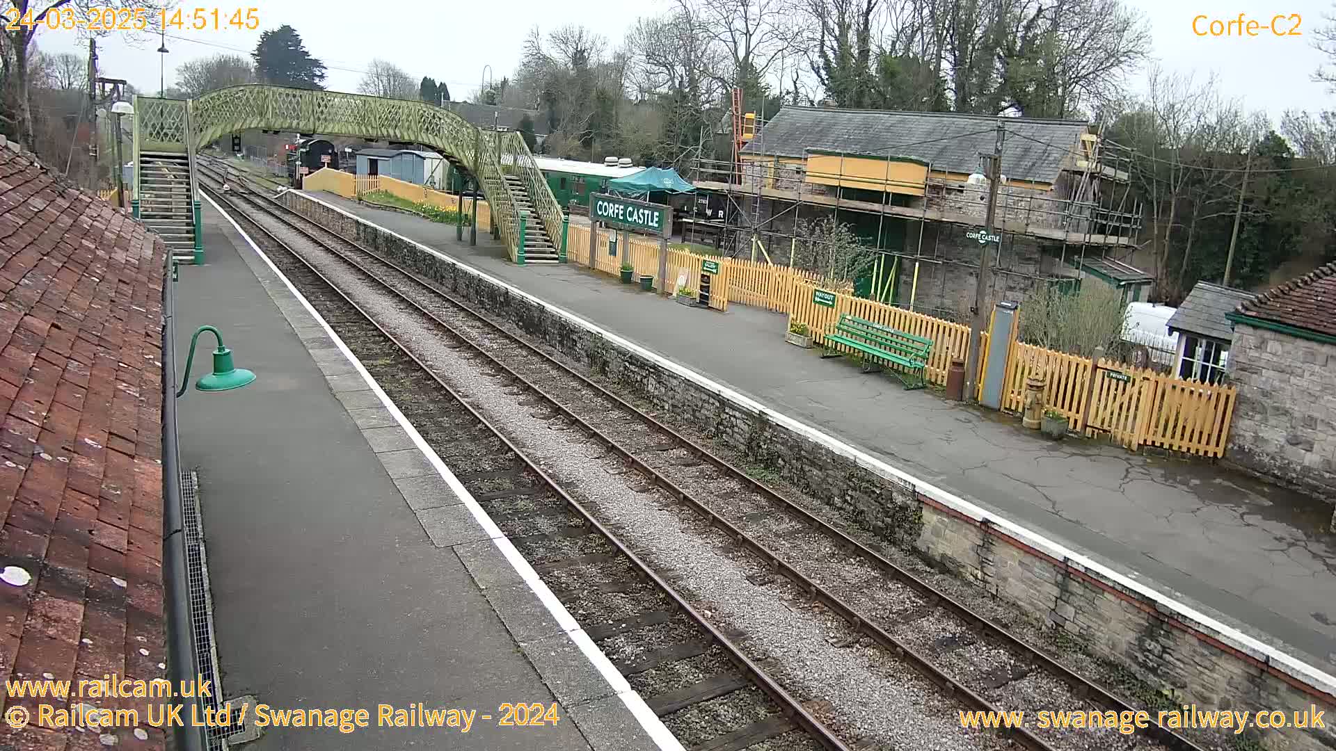 A train station platform with railway tracks, a pedestrian bridge, and buildings under construction on an overcast day.
