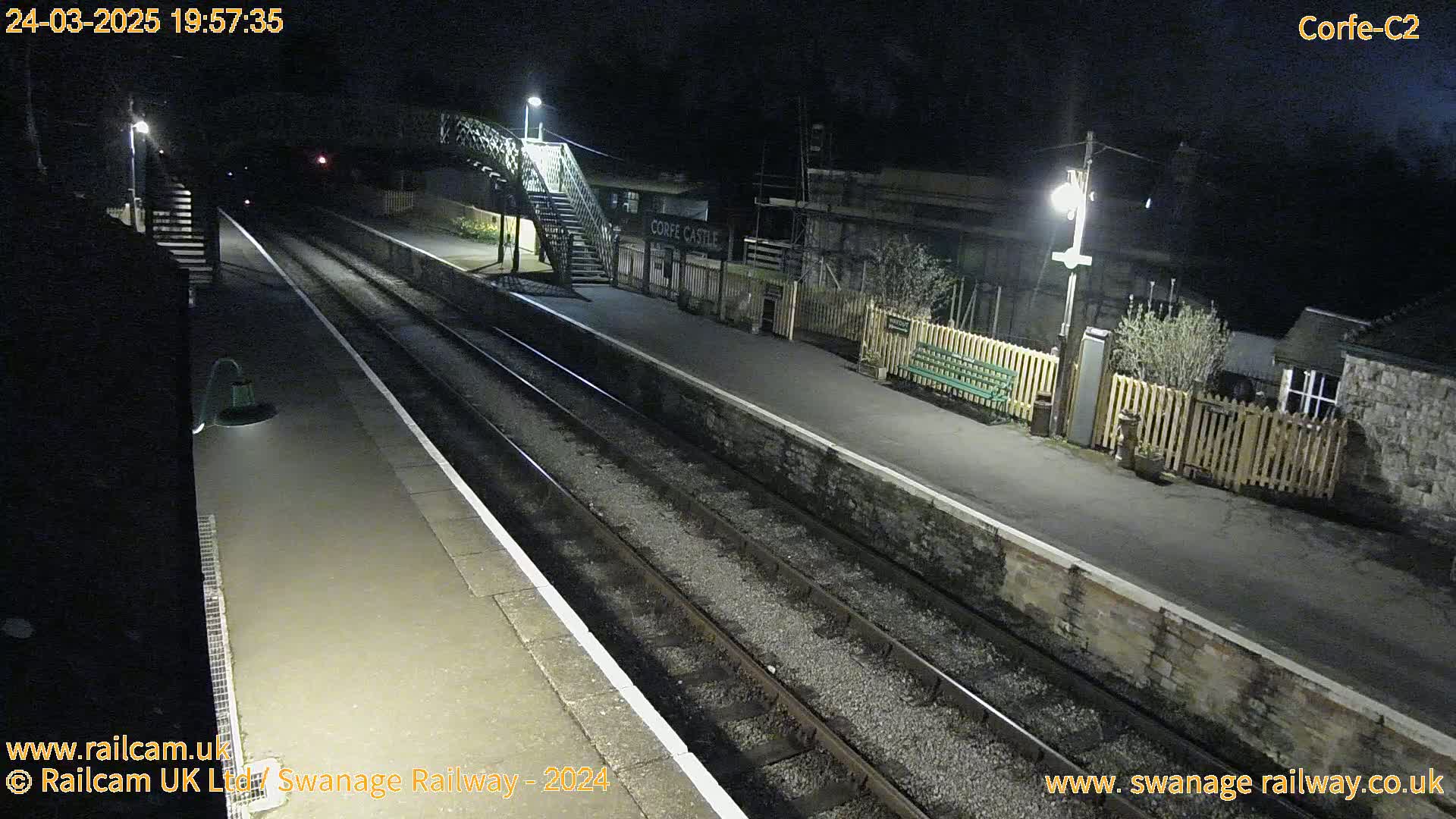 A nighttime view of a deserted train platform with two sets of tracks, lit by overhead lamps.