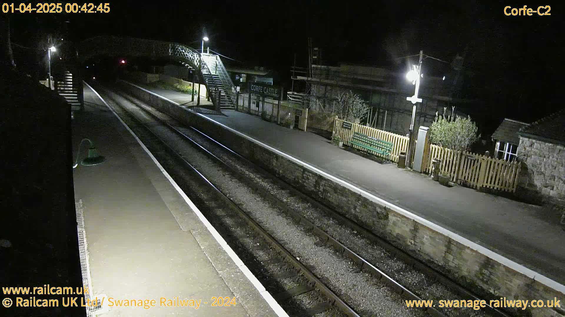 A deserted train platform at night, illuminated by artificial lights, with train tracks extending into the darkness.