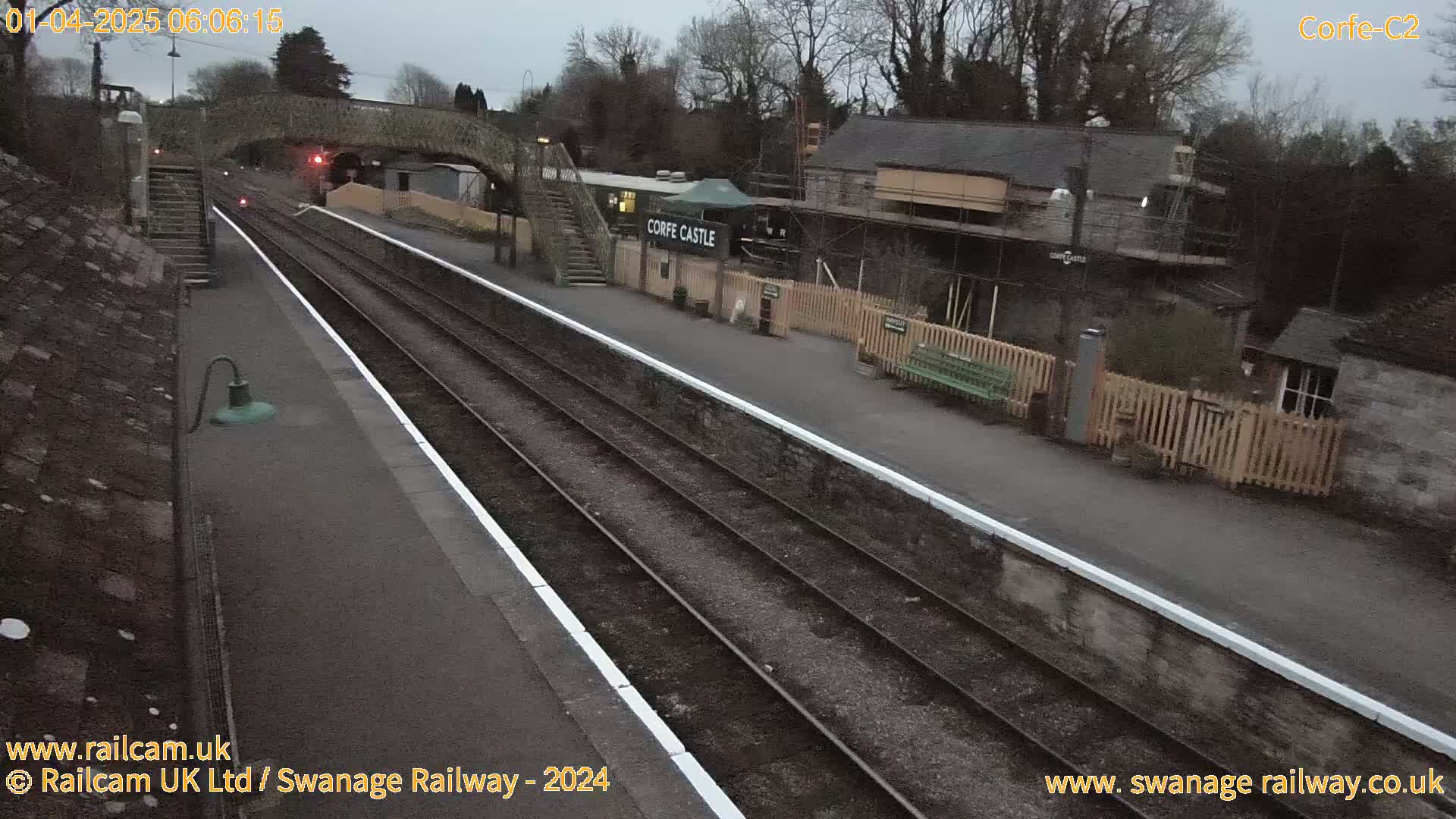 A train station platform with two sets of tracks under an overcast sky, featuring a pedestrian bridge, several buildings, and a wooden fence.