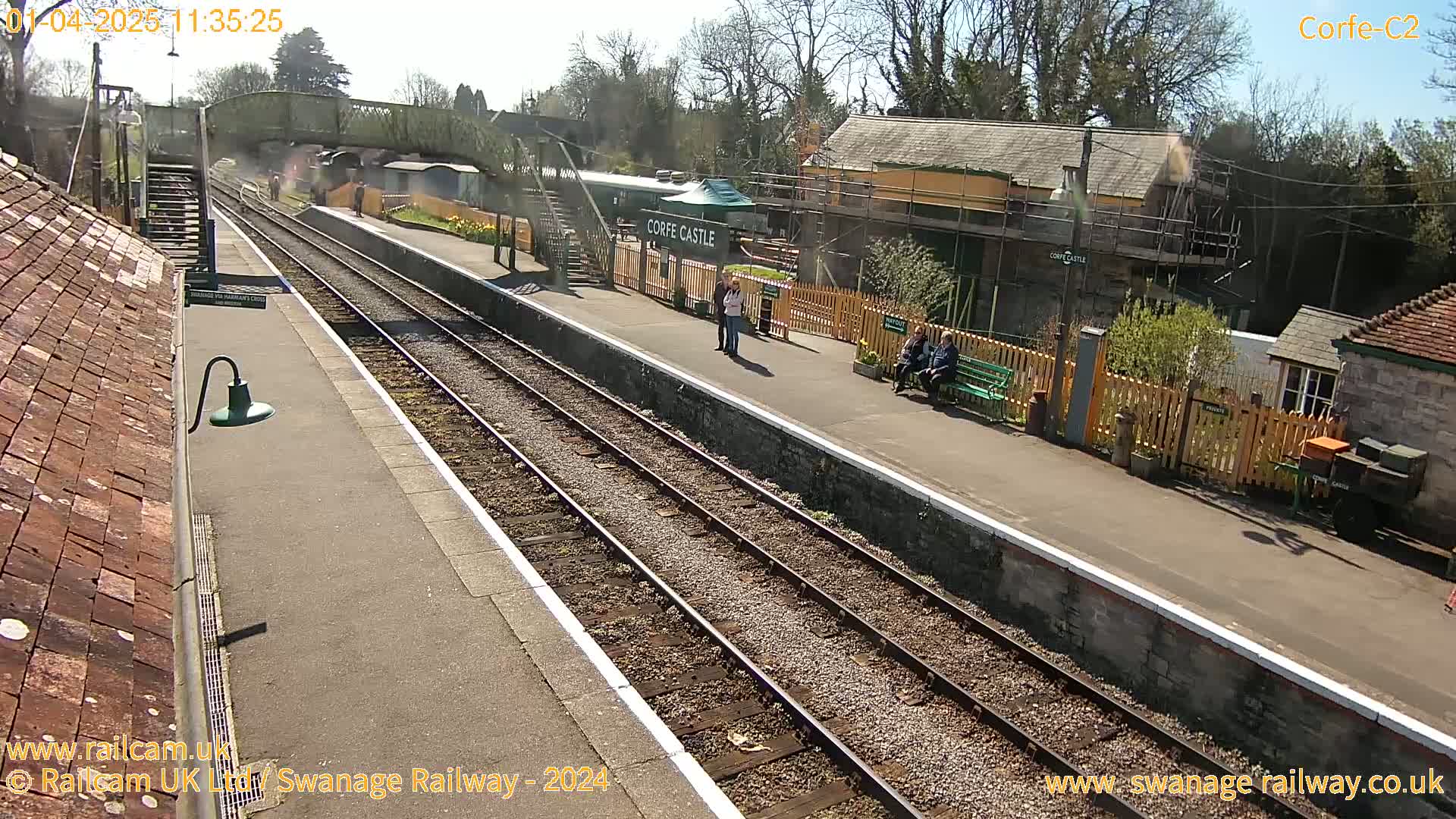 A sunny view of a train station platform with two sets of train tracks, several people, and station buildings.