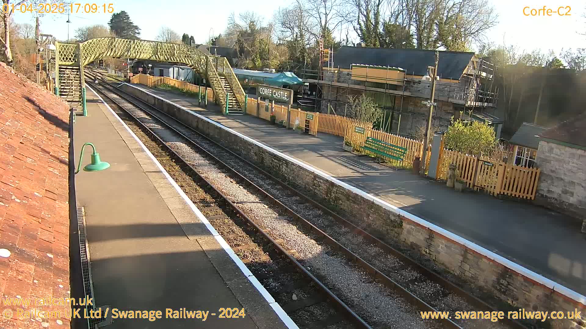 A train station platform with a railway line, a pedestrian bridge, and several buildings under a sunny sky.