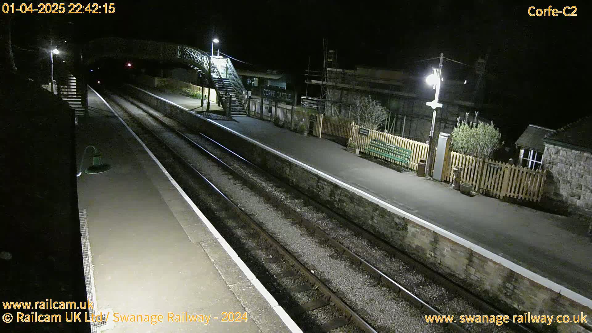 A nighttime view of a deserted train platform with two sets of tracks, a pedestrian bridge, and a station building.