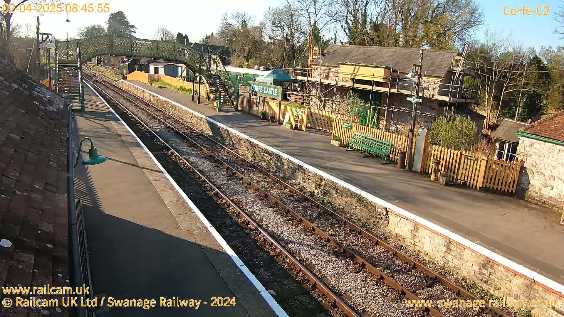 A sunny view of a railway station platform with train tracks, buildings, and a footbridge.