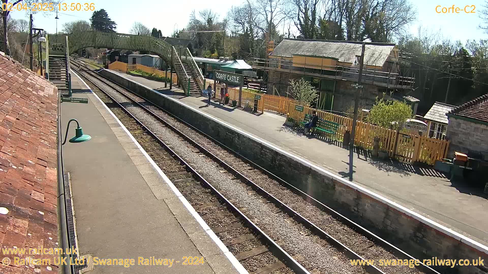A sunny day at a train station with two parallel train tracks, a platform, and several buildings, including a small station house and a footbridge.