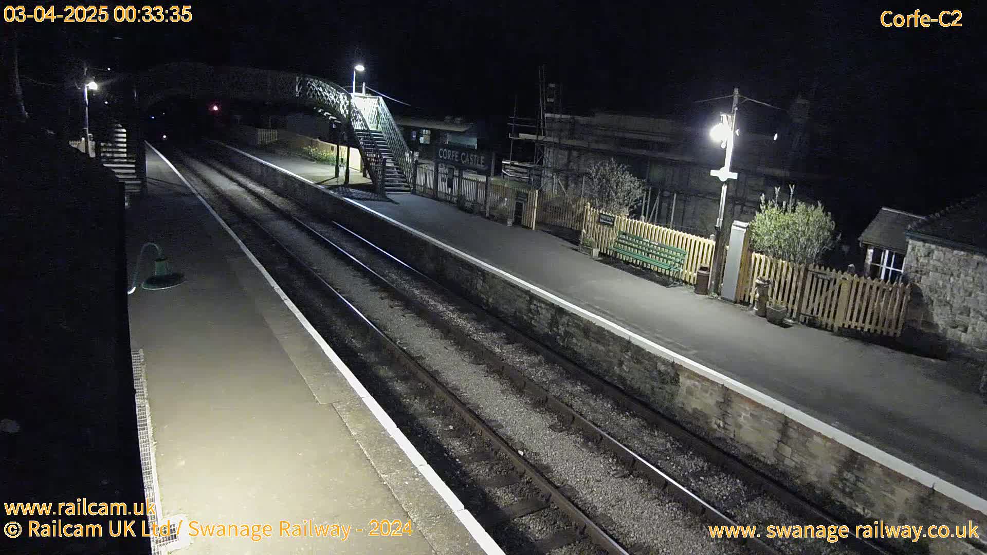A nighttime view of a deserted train platform with two sets of tracks, a pedestrian bridge, and a building.