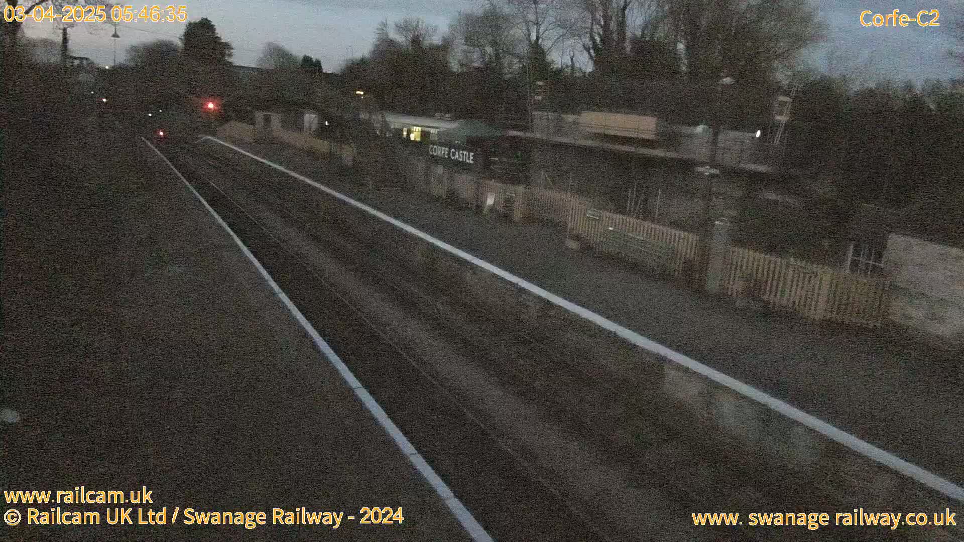 A nighttime view of a train station platform with two sets of tracks and a building labeled "Corfe Castle" in the background under a dark sky.