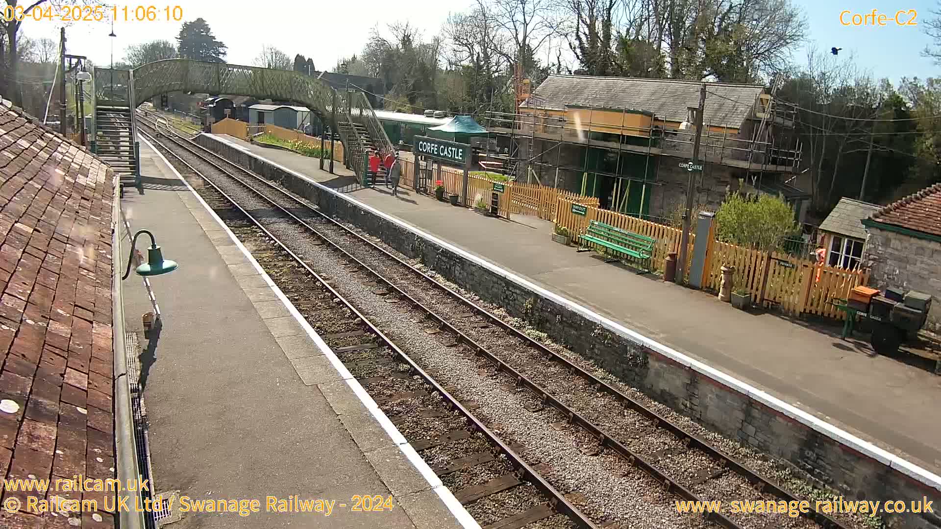 A sunny day at a train station with two parallel train tracks, a platform, and several buildings, including one under construction.