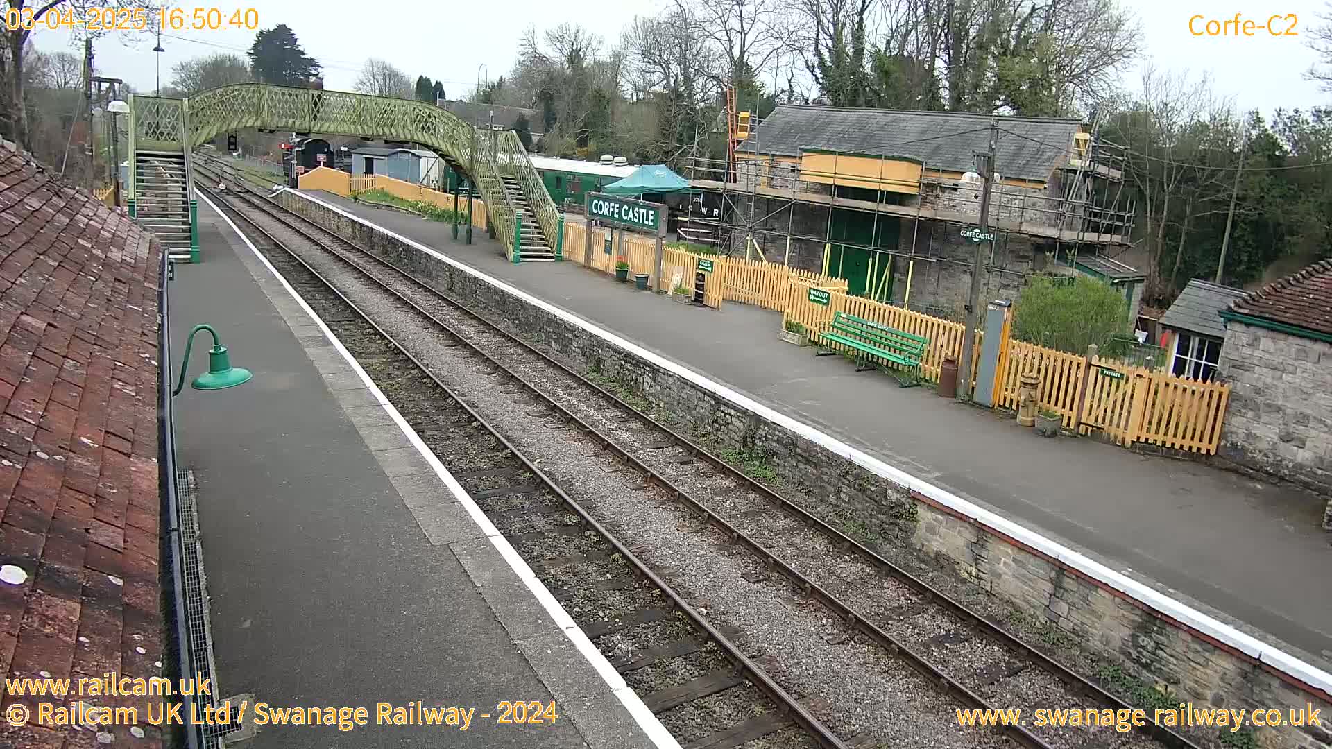 A train station with a footbridge, railway tracks, and several stone buildings under an overcast sky.