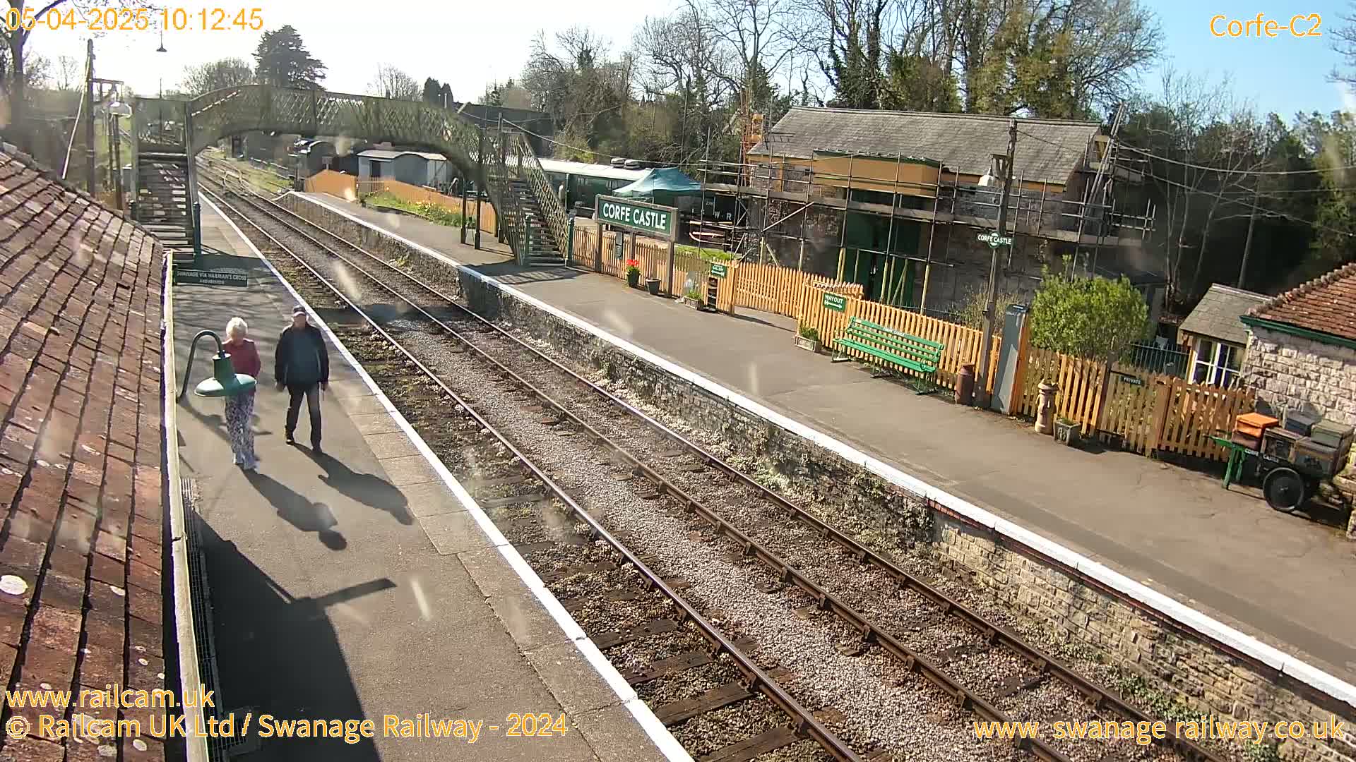 A sunny day at a train station with two people walking on the platform, railway tracks extending into the distance, and several buildings nearby.