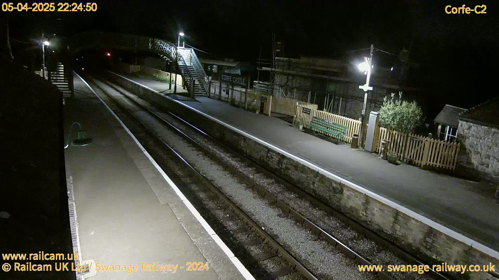 A nighttime view of a deserted train station platform with two sets of train tracks, a footbridge, and a building.