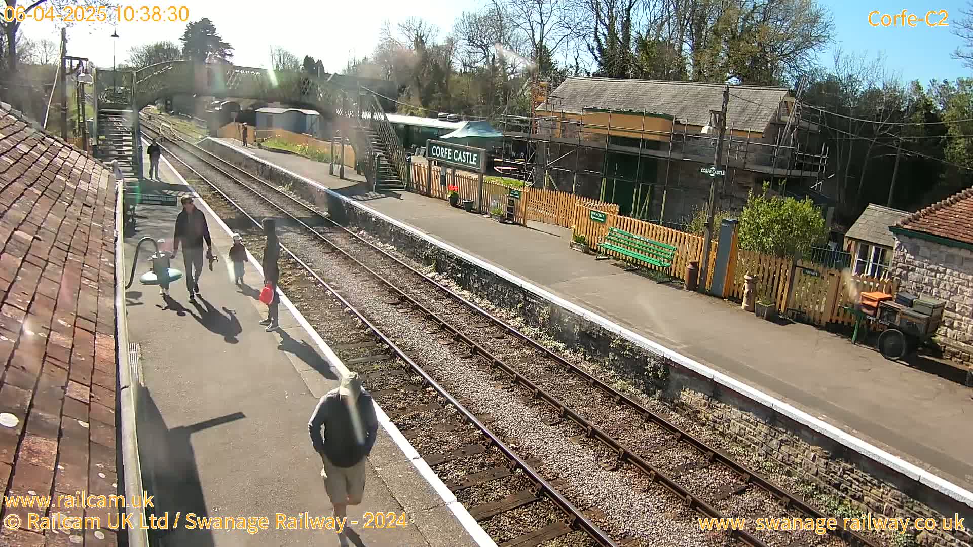 A sunny day at a train station shows a platform with several people walking, train tracks, and station buildings.