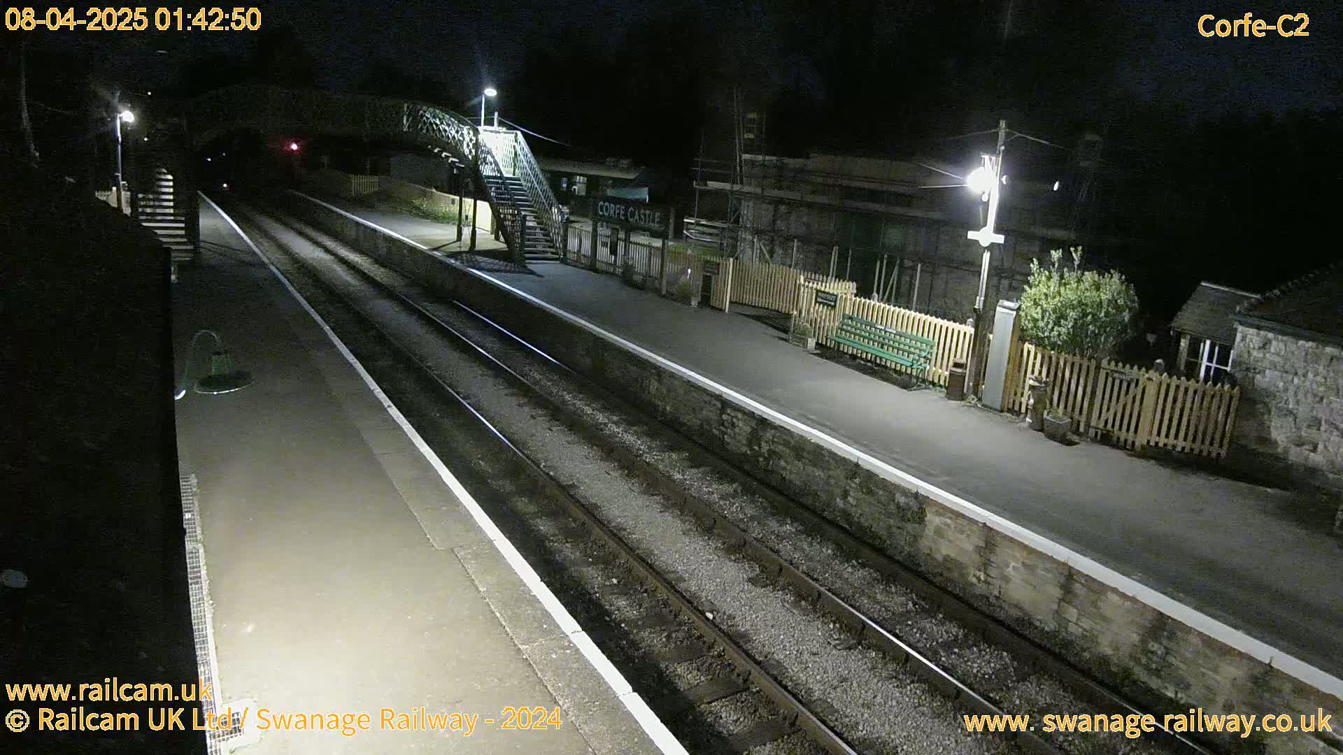 A nighttime view of a train station platform with two sets of train tracks, a footbridge, and a small building.