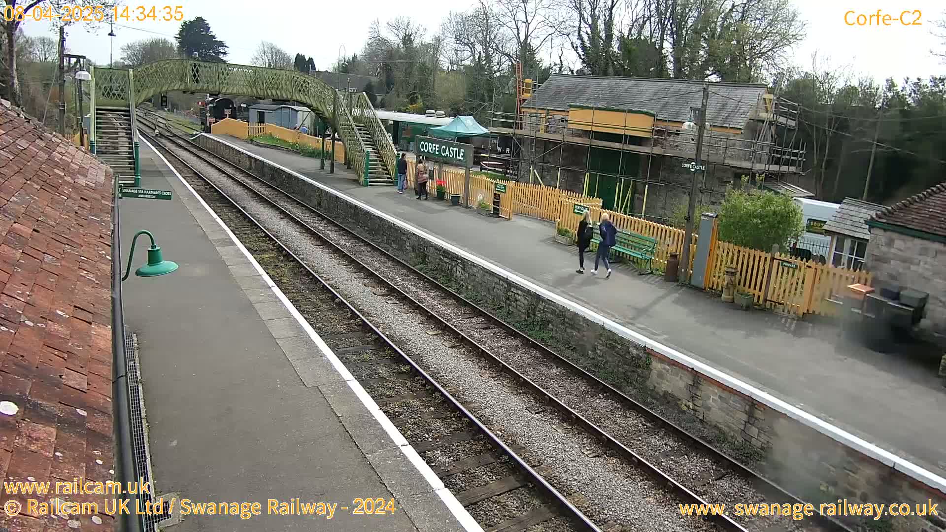 A railway station platform with two sets of train tracks, a pedestrian bridge, and several people walking around on a partly cloudy day.