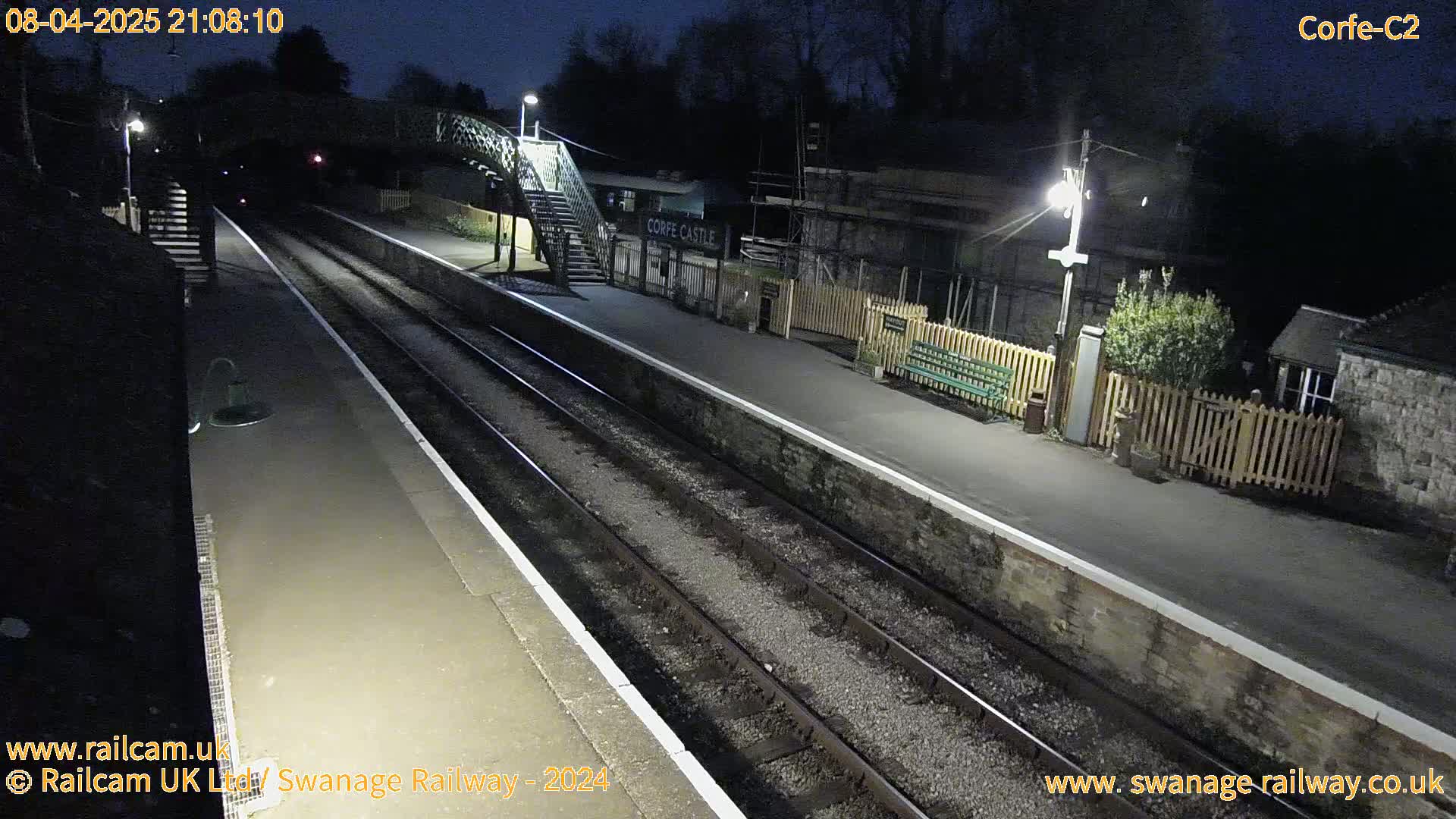 A nighttime view of a deserted train station with two train tracks, a footbridge, and platform lights illuminating the scene under a dark sky.