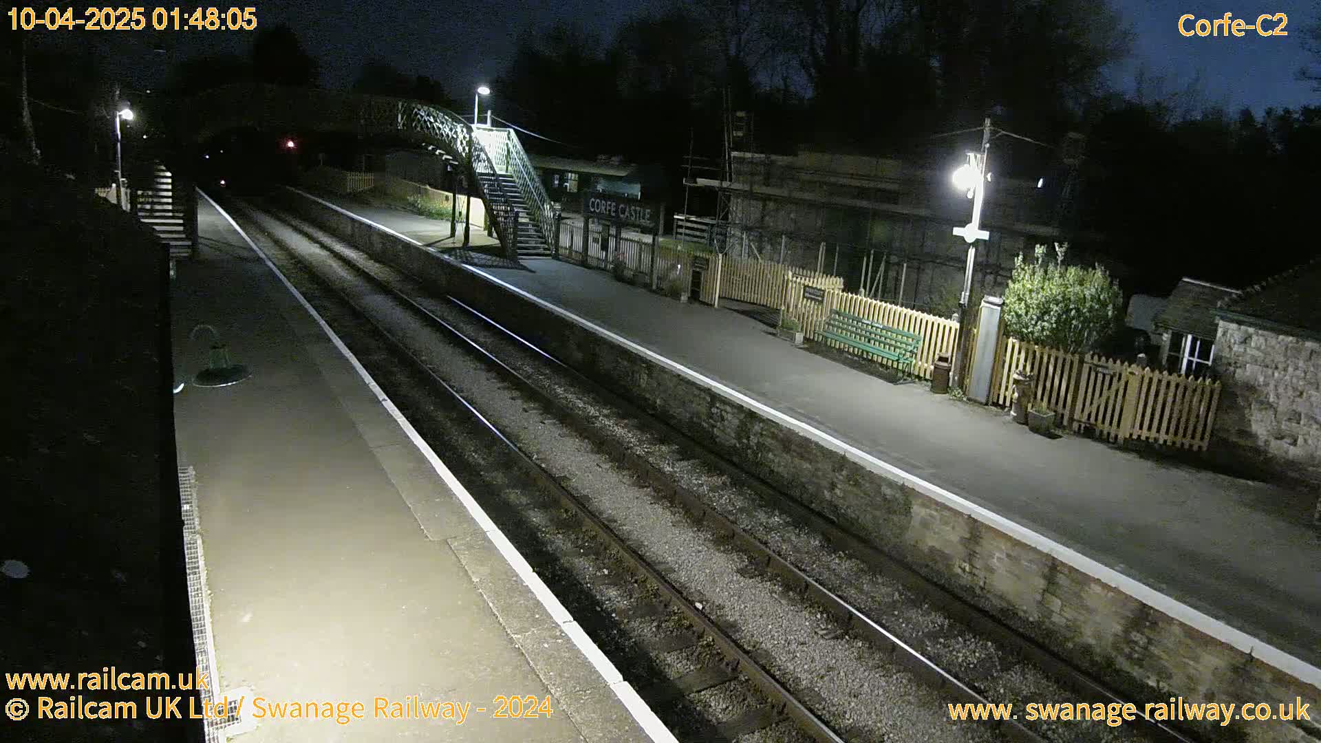 A nighttime view of a deserted train station platform with two train tracks, lit by several lamps, under a dark sky.