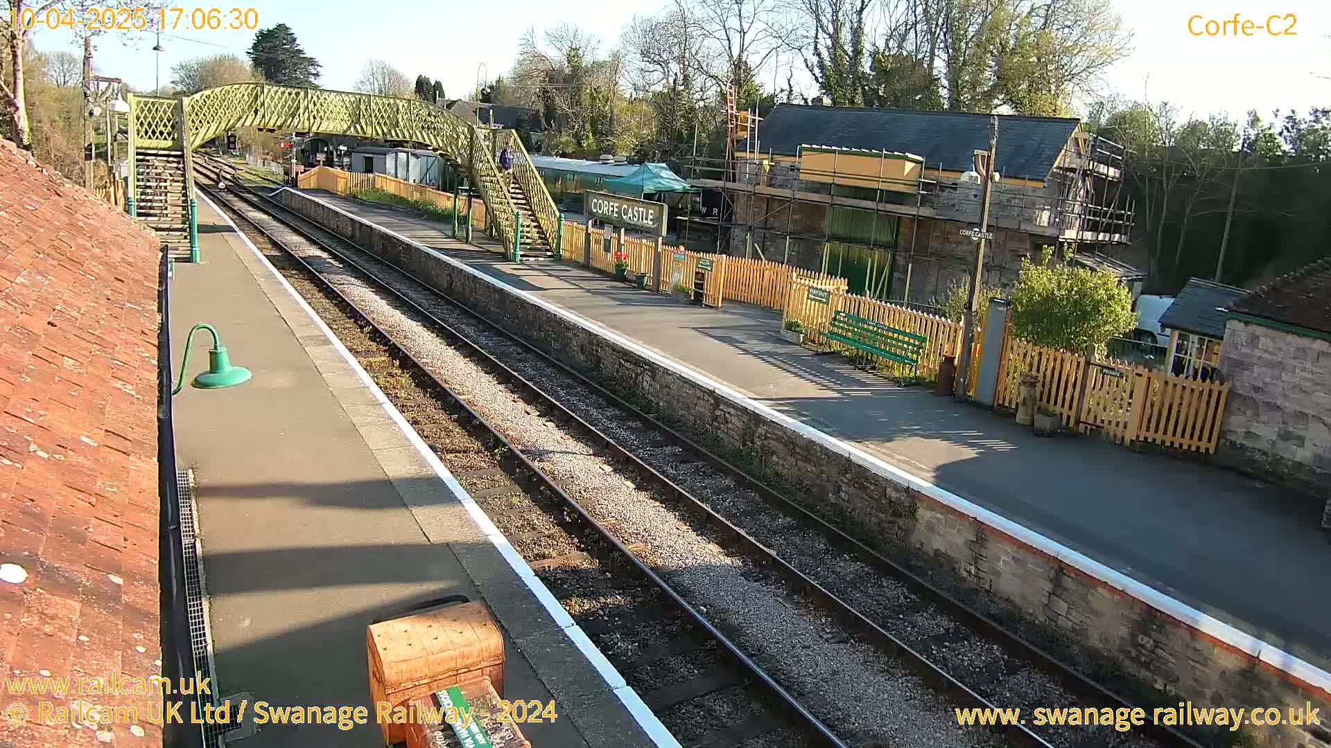 A sunny view of a train station platform with two parallel train tracks, a footbridge, and buildings undergoing construction.