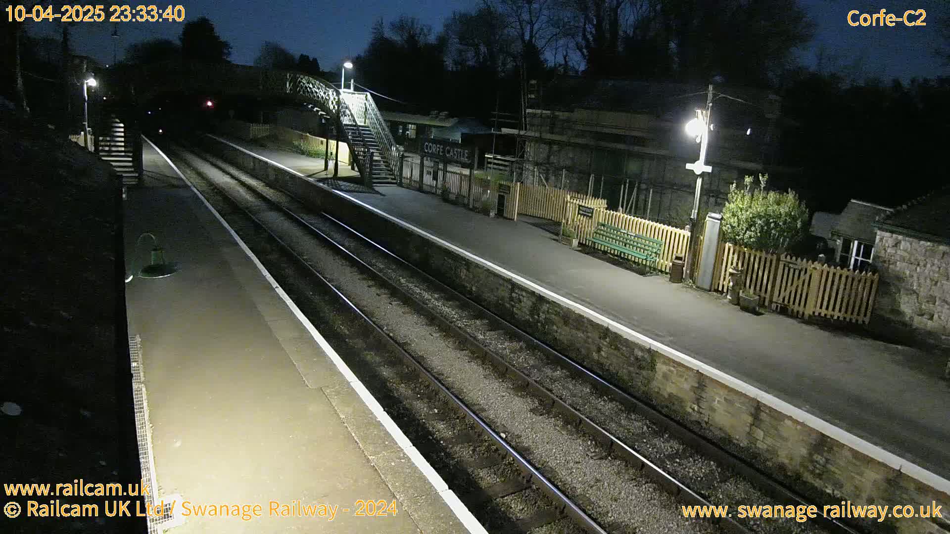 A nighttime view of a deserted train station platform with two sets of tracks under artificial lighting.