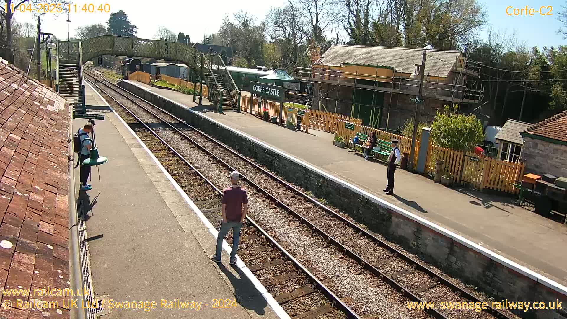 A sunny day at a train station shows several people standing near train tracks and buildings.