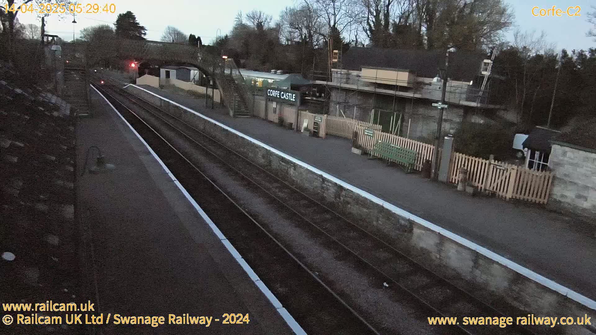 A train station platform with train tracks and a low stone wall under a twilight sky.