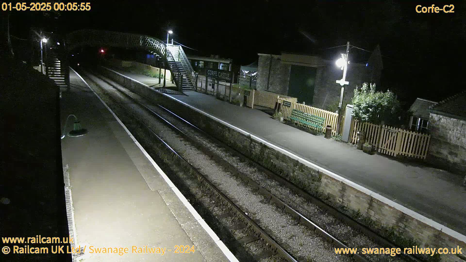 A nighttime view of a deserted train station platform with two sets of tracks, a footbridge, and a building.