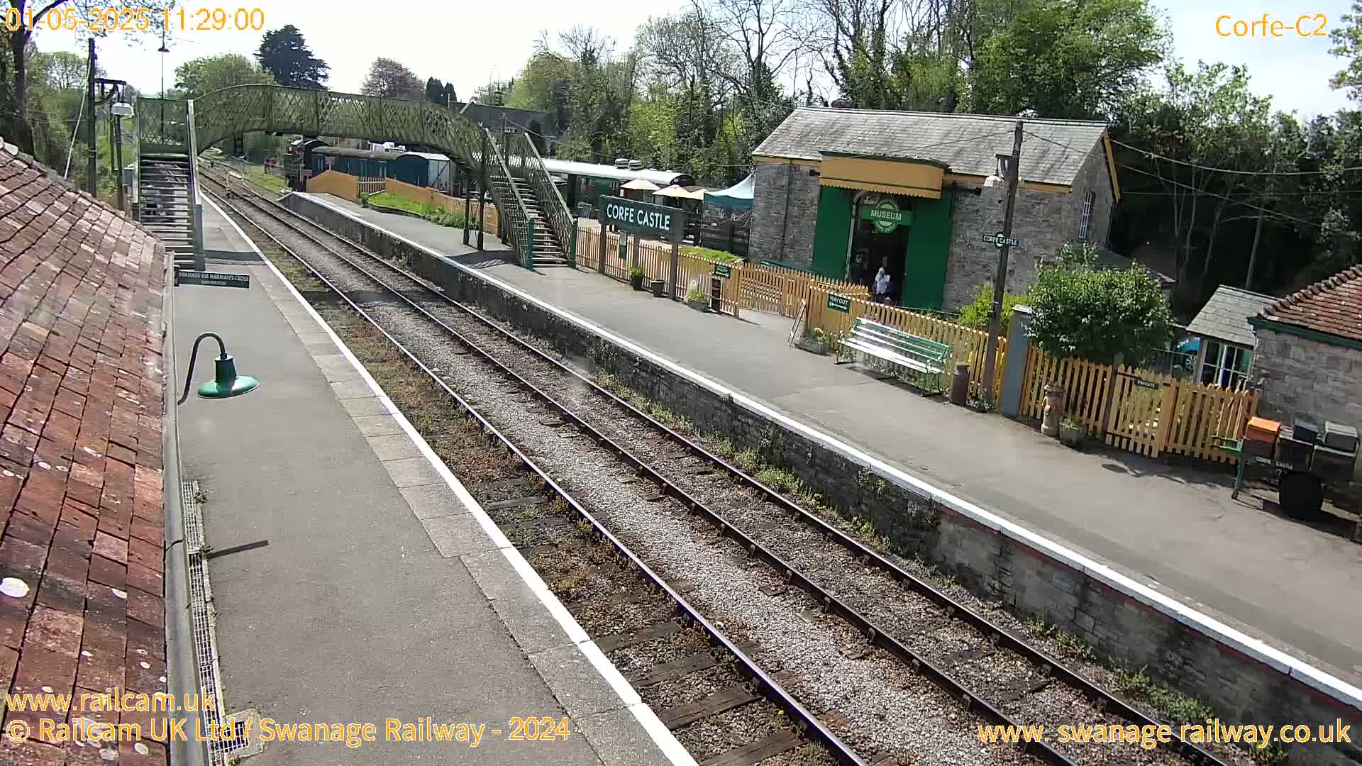 A sunny view of a train station with two parallel tracks, a footbridge, and several buildings including a museum.