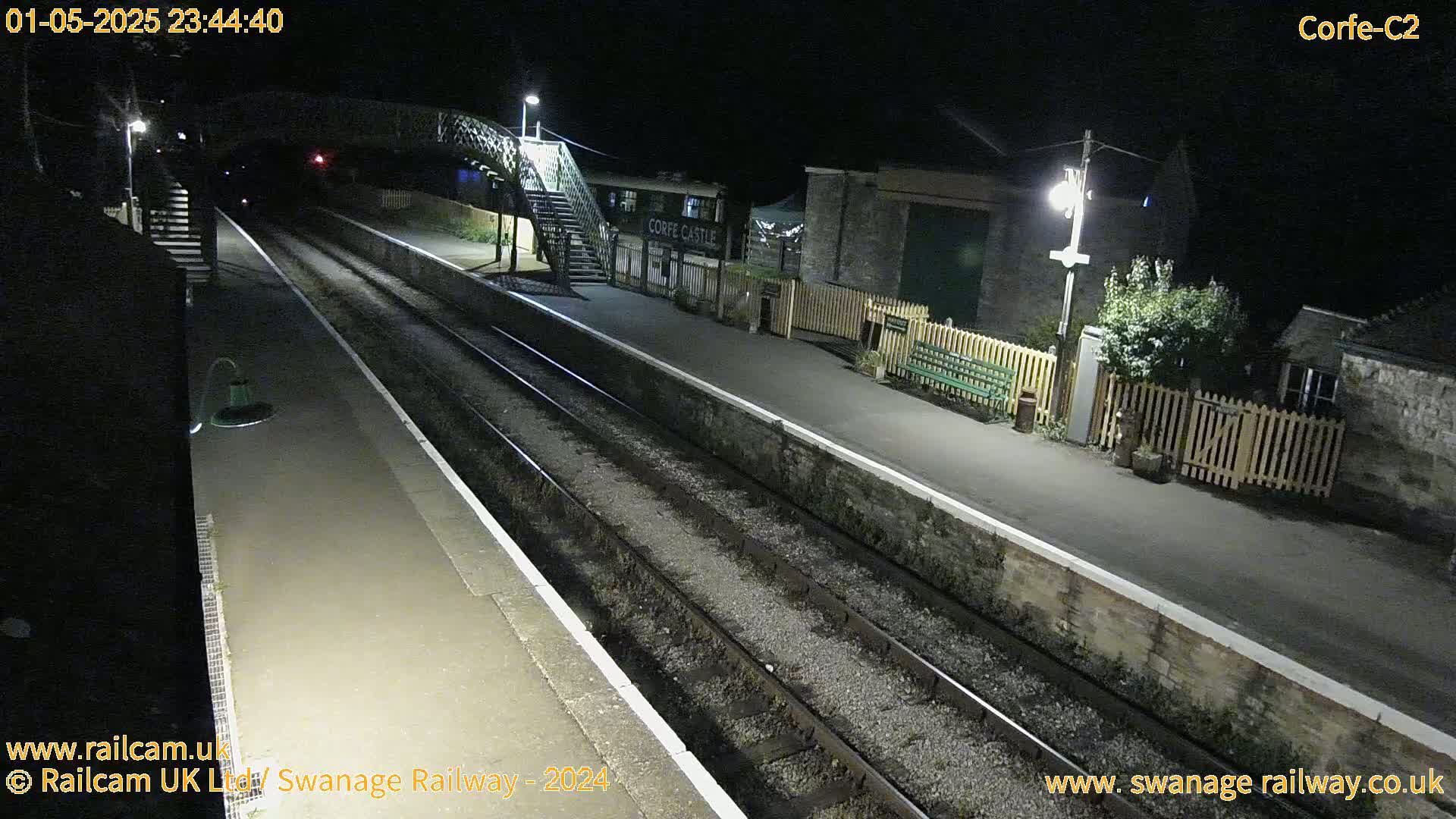 A nighttime view of a deserted train station platform with two sets of train tracks, a pedestrian bridge, and some station buildings illuminated by artificial light.