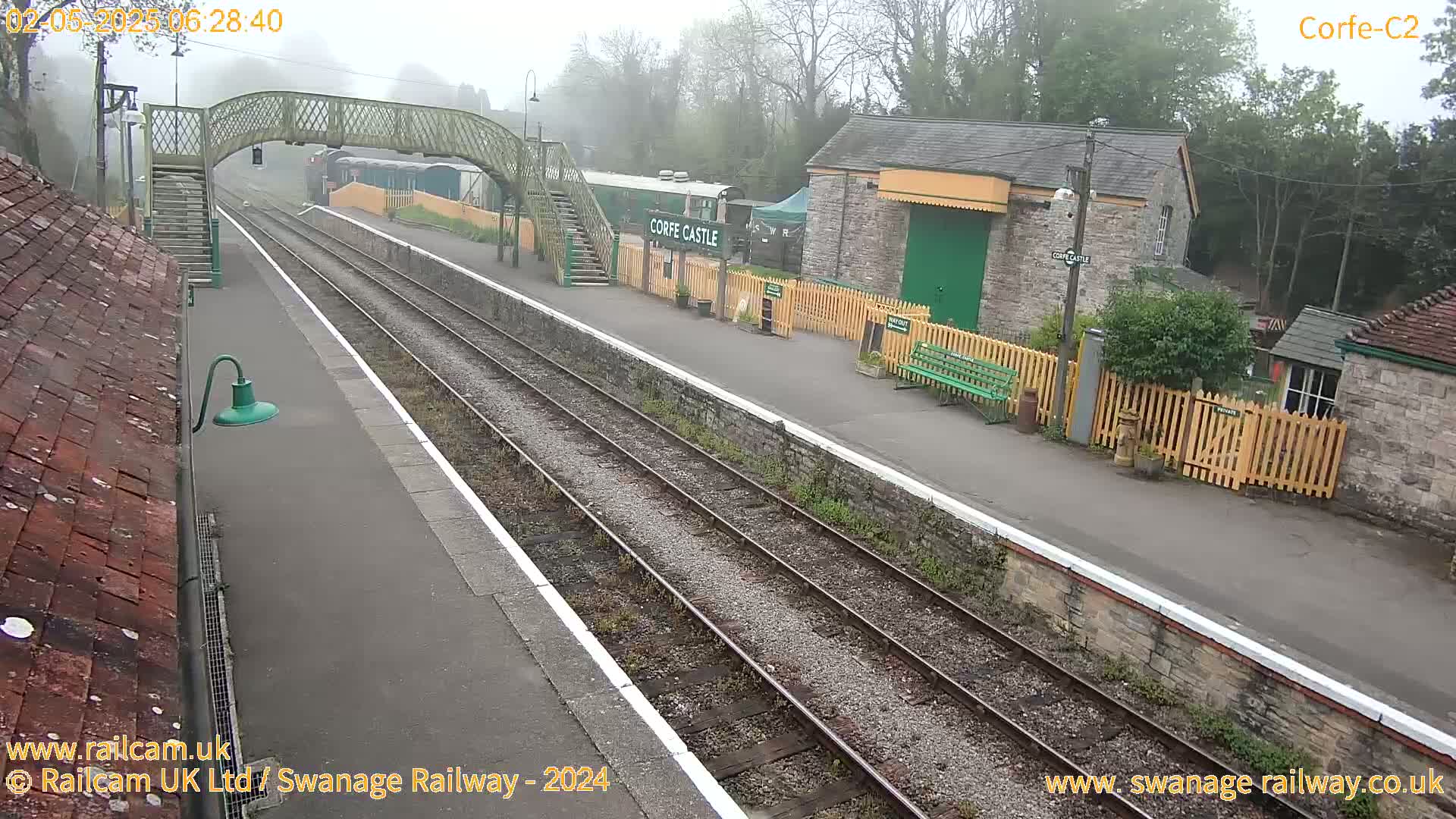 A misty view of a train station platform with railway tracks, a pedestrian bridge, and station buildings.