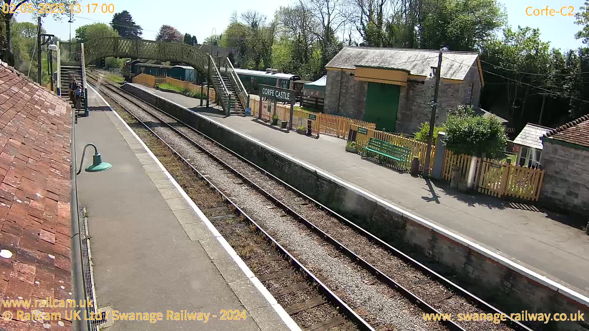 A sunny day at a train station with a railway track, platform, buildings, and a footbridge.